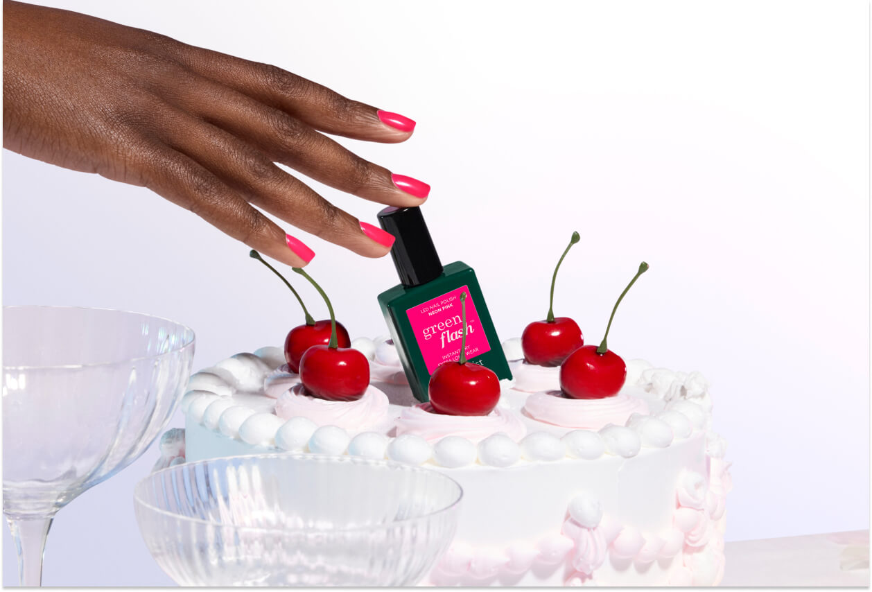 A hand with pink nail polish places a green nail polish bottle labeled green / flee among cherries on a white frosted cake, with empty glass bowls nearby.