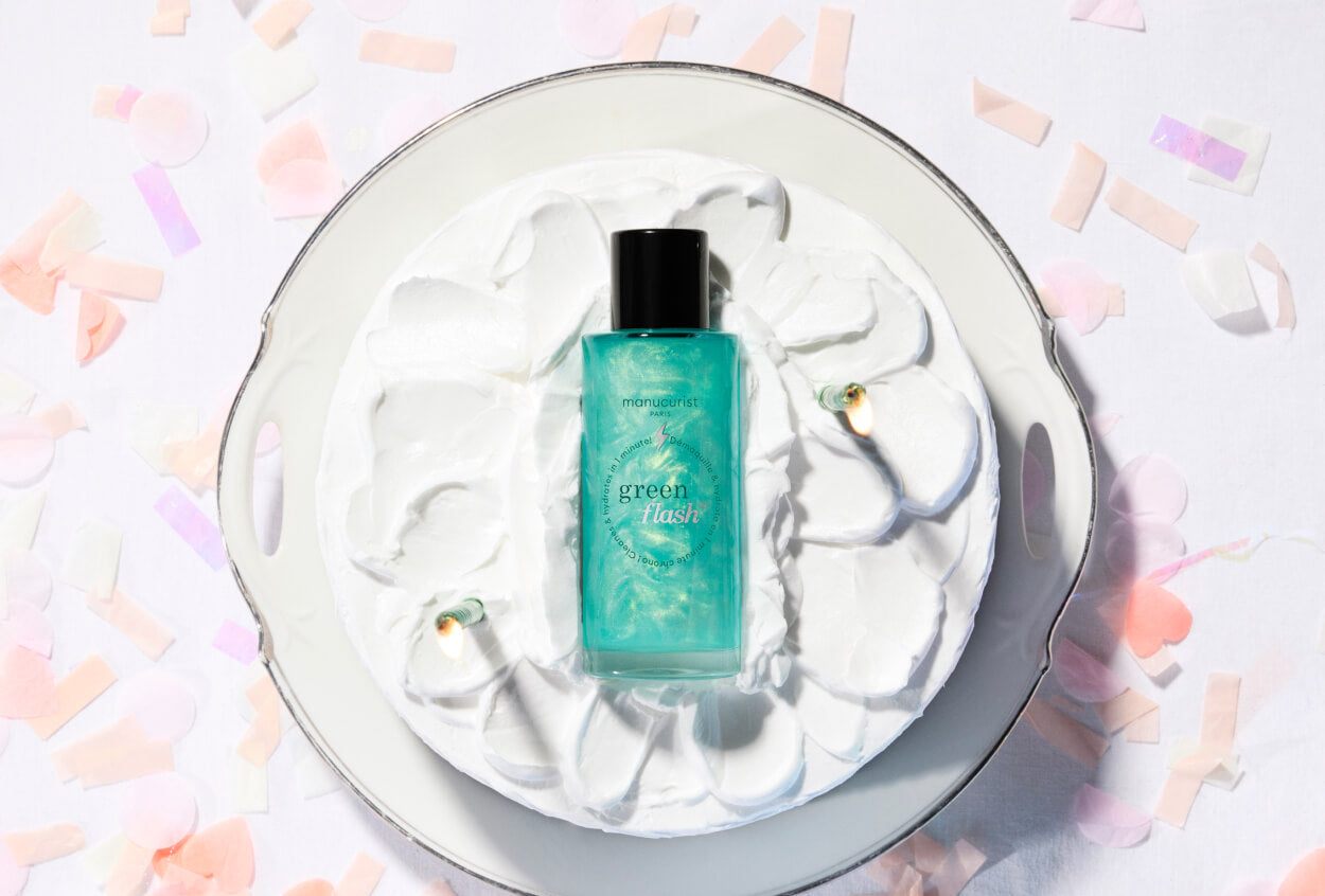 A turquoise bottle of “green flask” perfume stands on a plate topped with swirls of white whipped cream, surrounded by pastel confetti on a white background.
