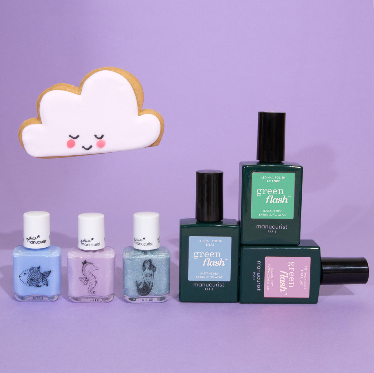 Five bottles from the Manucurist You Me Under the Sea Green Flash™ Set are arranged on a purple surface. Three pastel shades with illustrated animal labels are featured, with a smiling cloud-shaped cookie in the background.