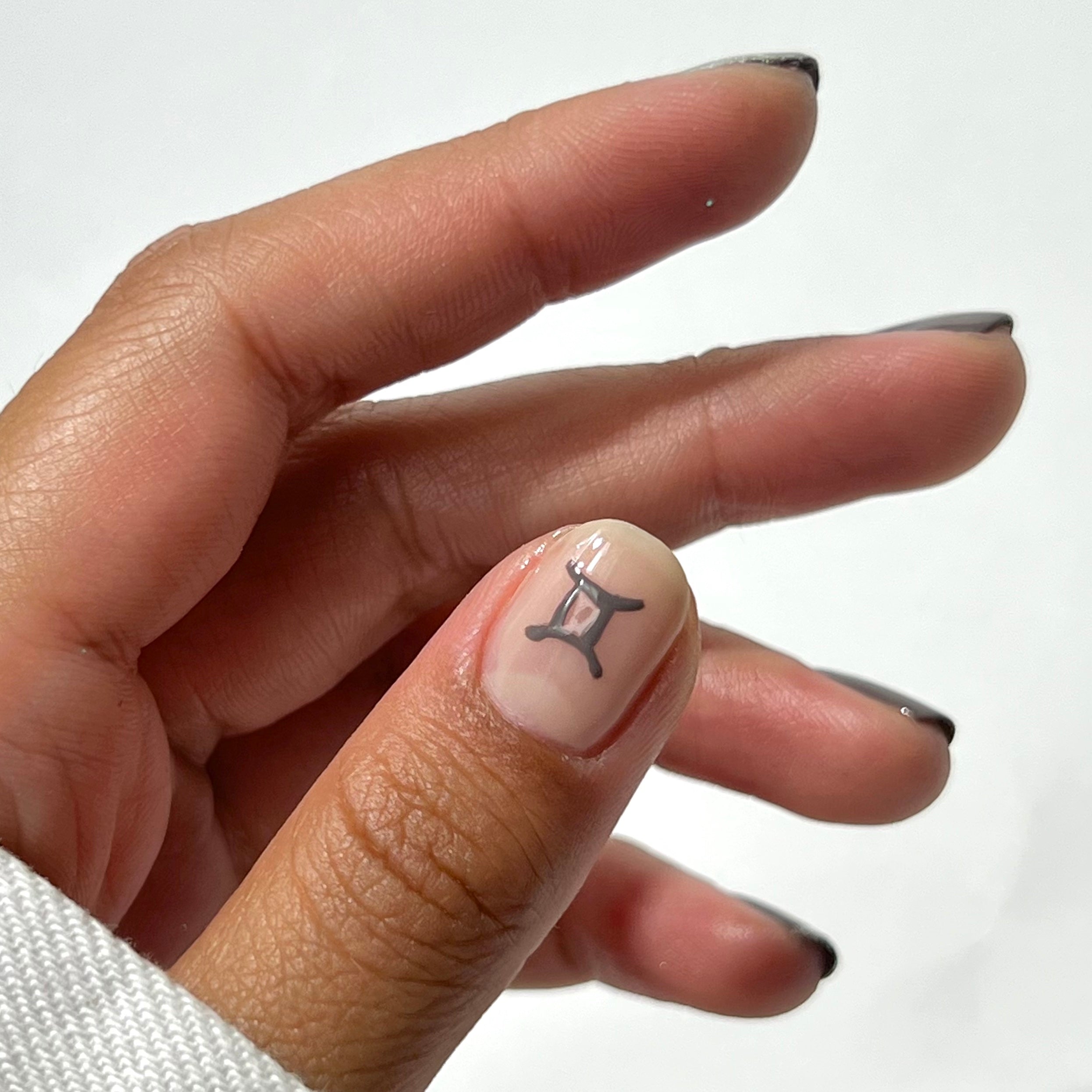A hand with short, neatly manicured nails, featuring a black Gemini zodiac symbol on the thumbnail. The nails have a clear base with black tips, and the background is plain white.