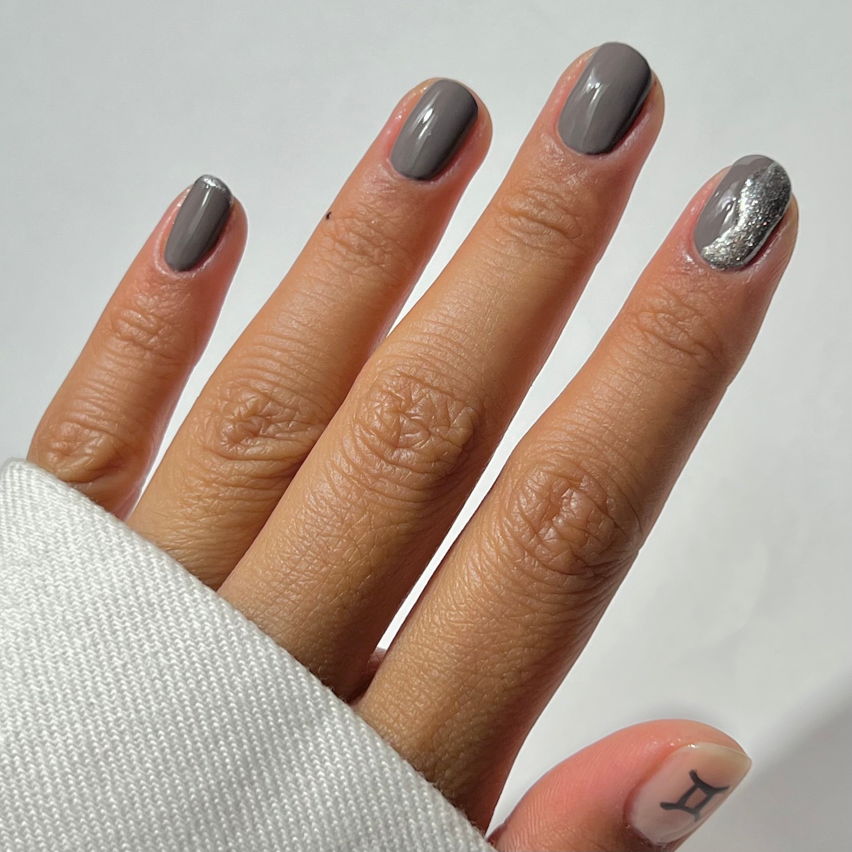 A hand with short, neatly manicured nails painted in gray polish; the ring finger features a silver glitter accent. The thumb has a small Gemini zodiac symbol. The person wears a white long-sleeve shirt.