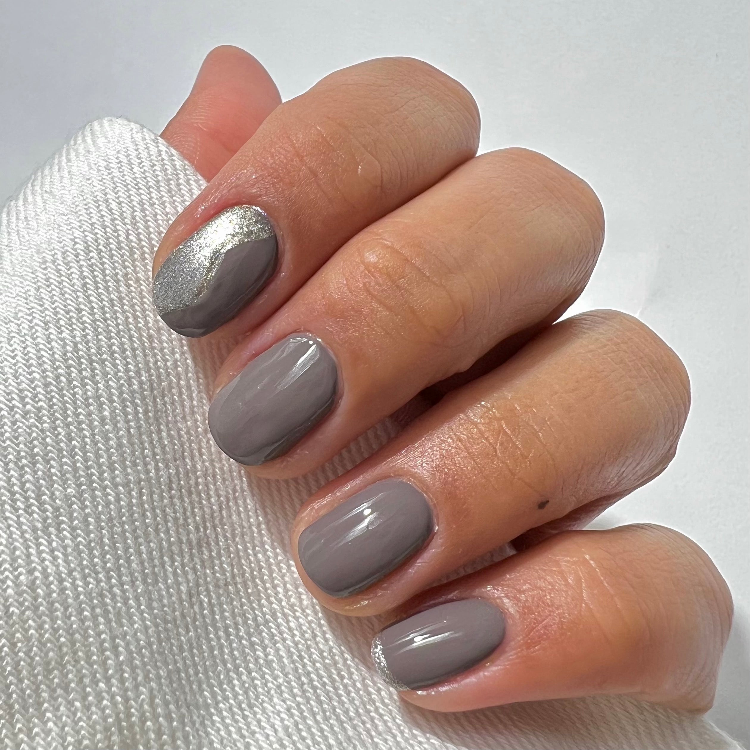 A hand with neatly manicured nails painted in glossy gray polish; the index fingernail features a silver wavy accent near the cuticle. The hand rests on a white knitted fabric.