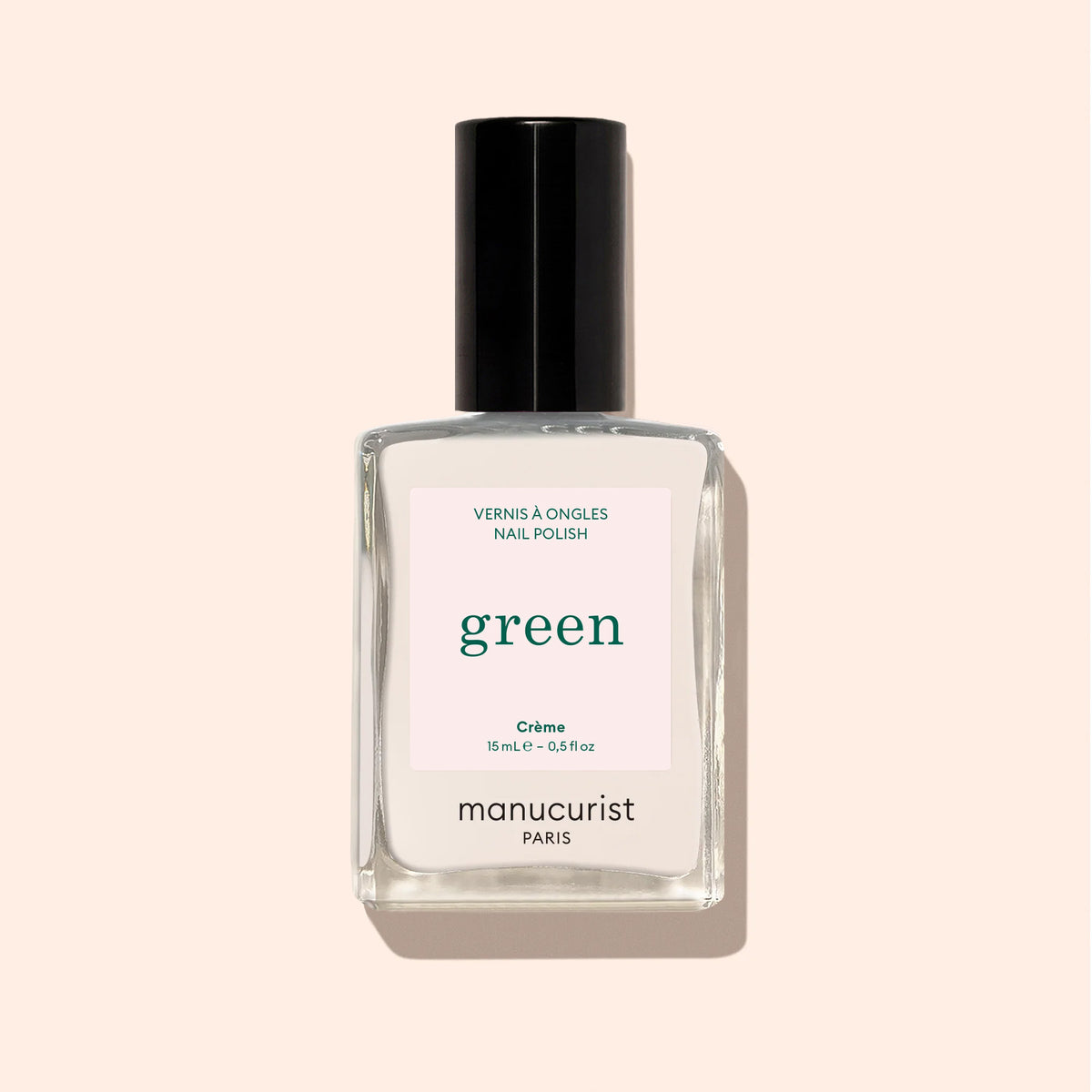 Un flacon en verre transparent de vernis à ongles Manucurist Crème, avec un bouchon noir et une étiquette rose clair, repose sur un fond beige.