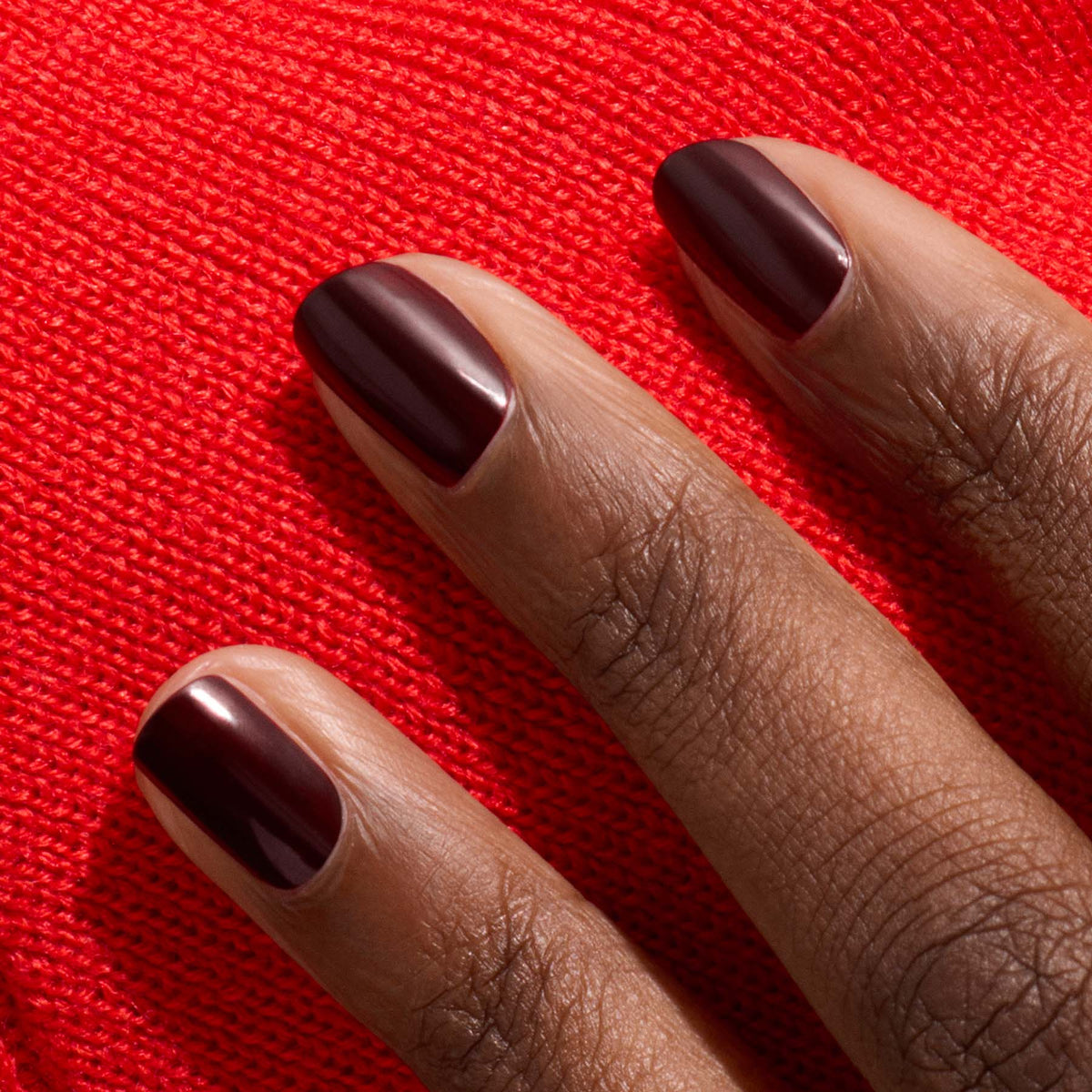 Trois ongles courts parfaitement manucurés avec le vernis rouge profond Hollyhock de Manucurist reposent sur un tissu rouge éclatant, mettant en valeur une peau mate.