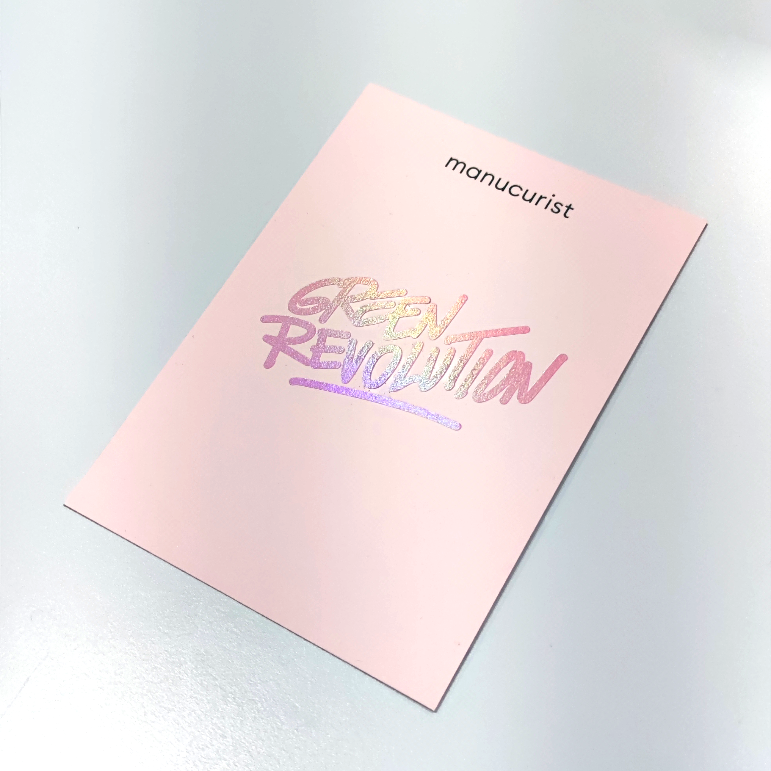 A pink card on a white surface with the word manucurist at the top and Green Revolution in shiny, iridescent lettering below.