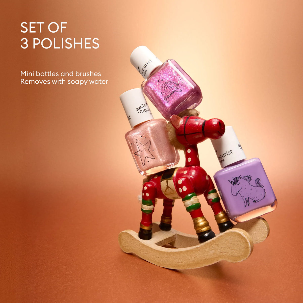 Three mini nail polish bottles from the Petite Manucurist Trio by Manucurist—in pink, rose gold, and lavender—are balanced on a small wooden rocking horse against a copper-toned background. Text reads: Petite Manucurist Trio. Includes 3 mini bottles and brushes. Children’s nail polish made with natural origin ingredients; washes off easily with soapy water—perfect for holiday gifting.