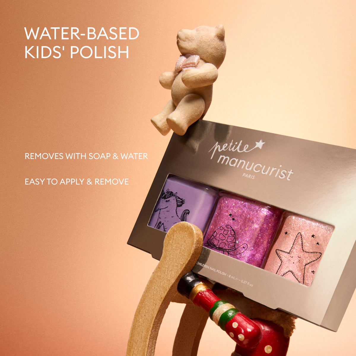 The Petite Manucurist Trio by Manucurist—a kids’ nail polish set featuring purple, pink, and glitter shades—is displayed on a rocking horse. Text reads: Water-based children’s nail polish of natural origin. Removes with soap water. Easy to apply and remove—perfect as a holiday gift. A teddy bear is seated above.