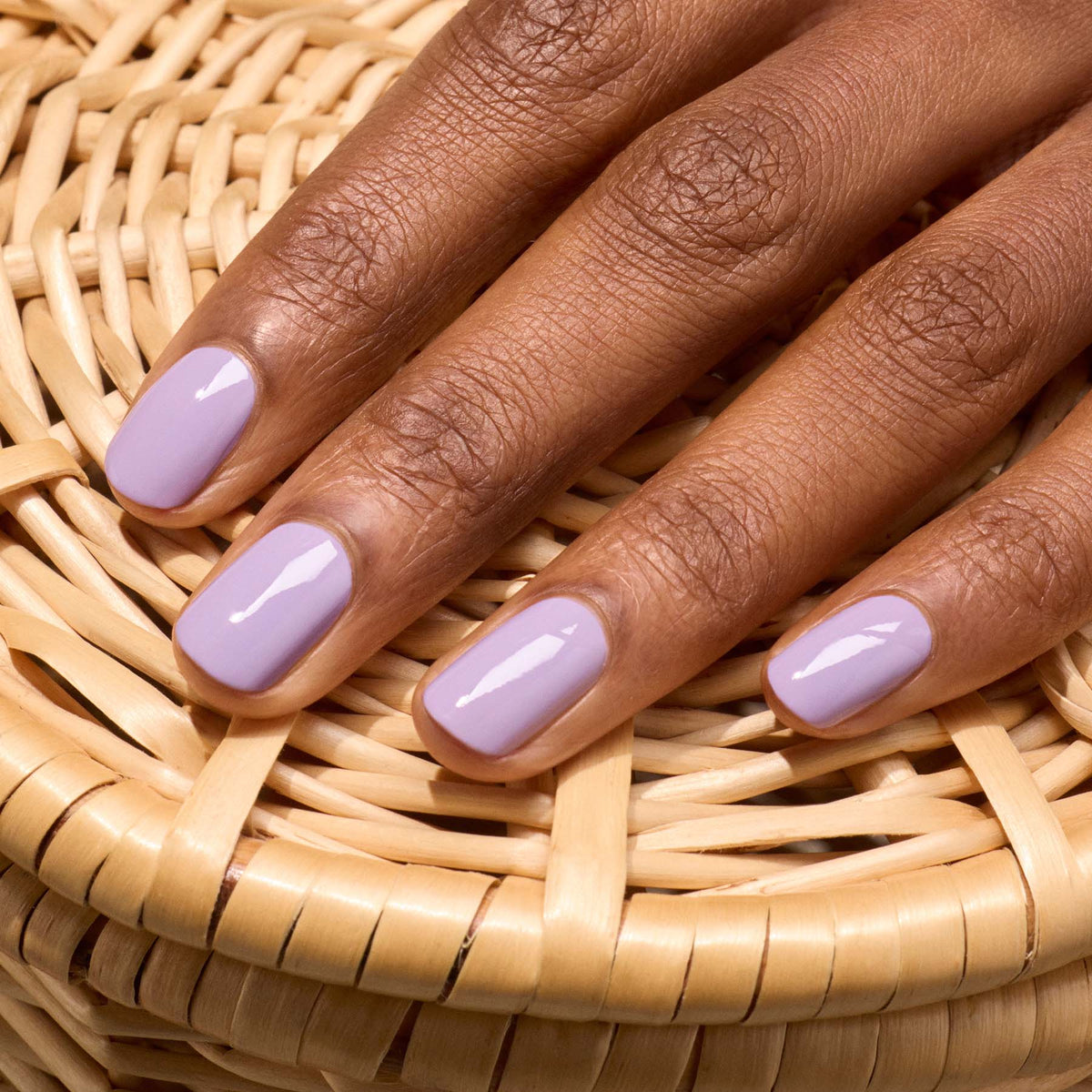 Gros plan d’une main aux ongles courts soigneusement manucurés, recouverts du vernis gel Lisa Lilas brillant de Manucurist, posée sur de l’osier et évoquant subtilement les années 90.