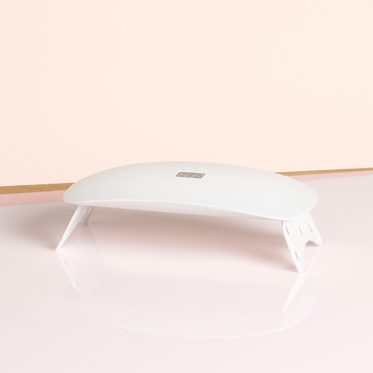 The Manucurist Green Flash™ 24W Lamp—a compact, white, curved LED lamp with four legs—sits on a glossy pink surface, ideal for Green Flash gel polish manicures, set against a beige and pink backdrop.
