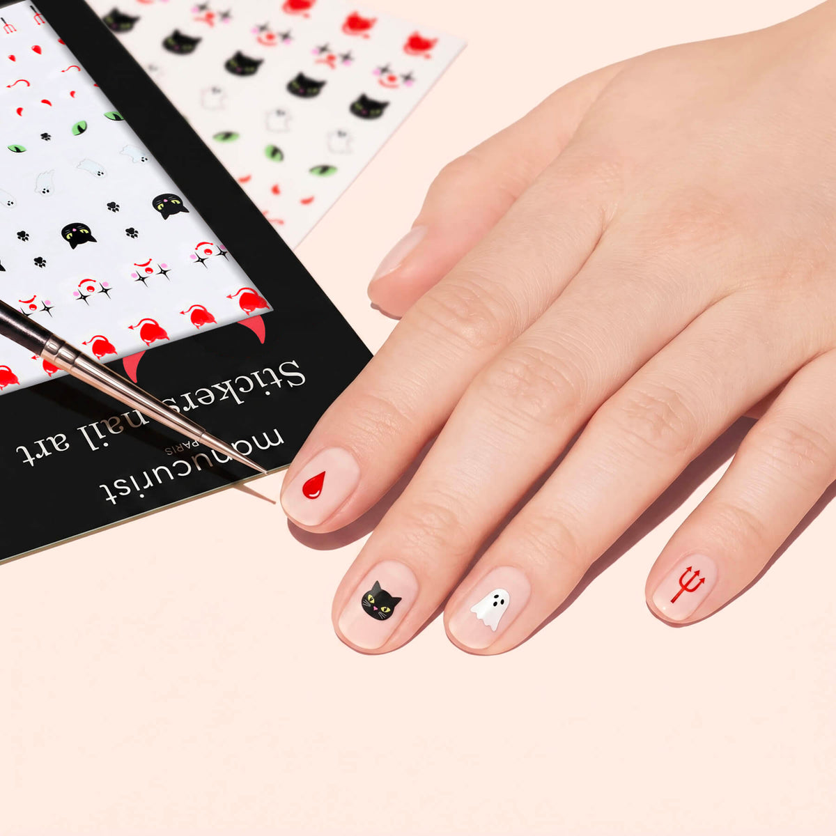 Une main ornée de stickers Nail Art Halloween Manucurist — cœur rouge, chat noir, fantôme et trident rouge — tandis qu’un pinceau pointe vers les ongles et qu’une planche de stickers aux motifs effrayants se trouve à proximité.