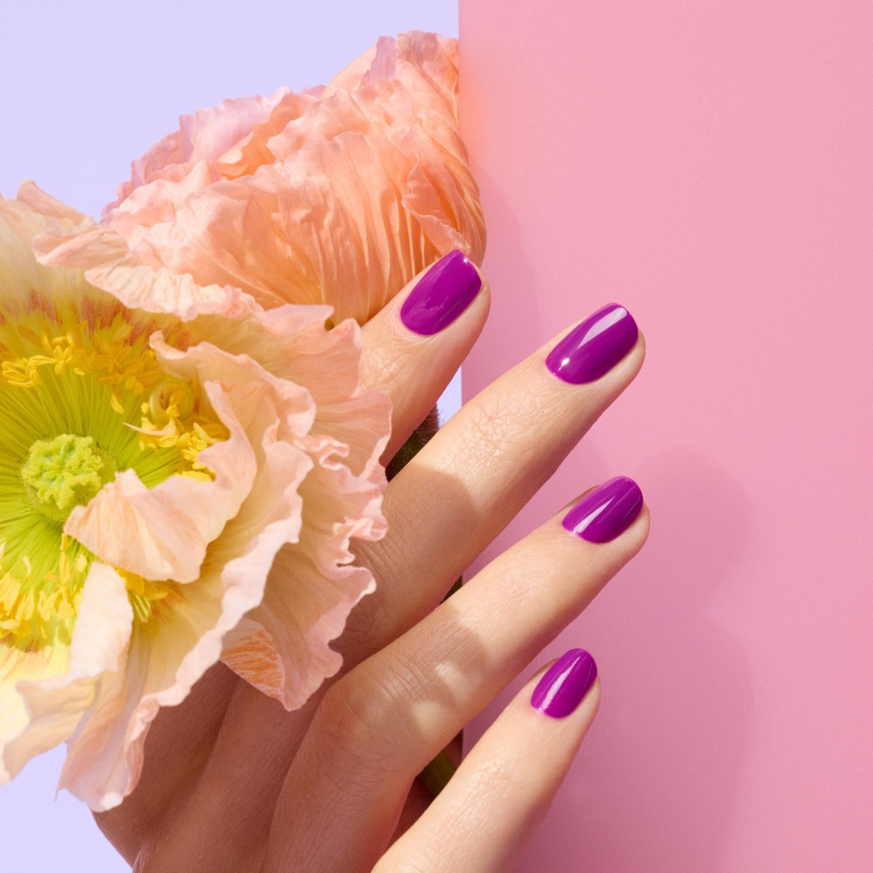 A hand with glossy purple nail polish holds peach-colored flowers against a pink and lavender background.