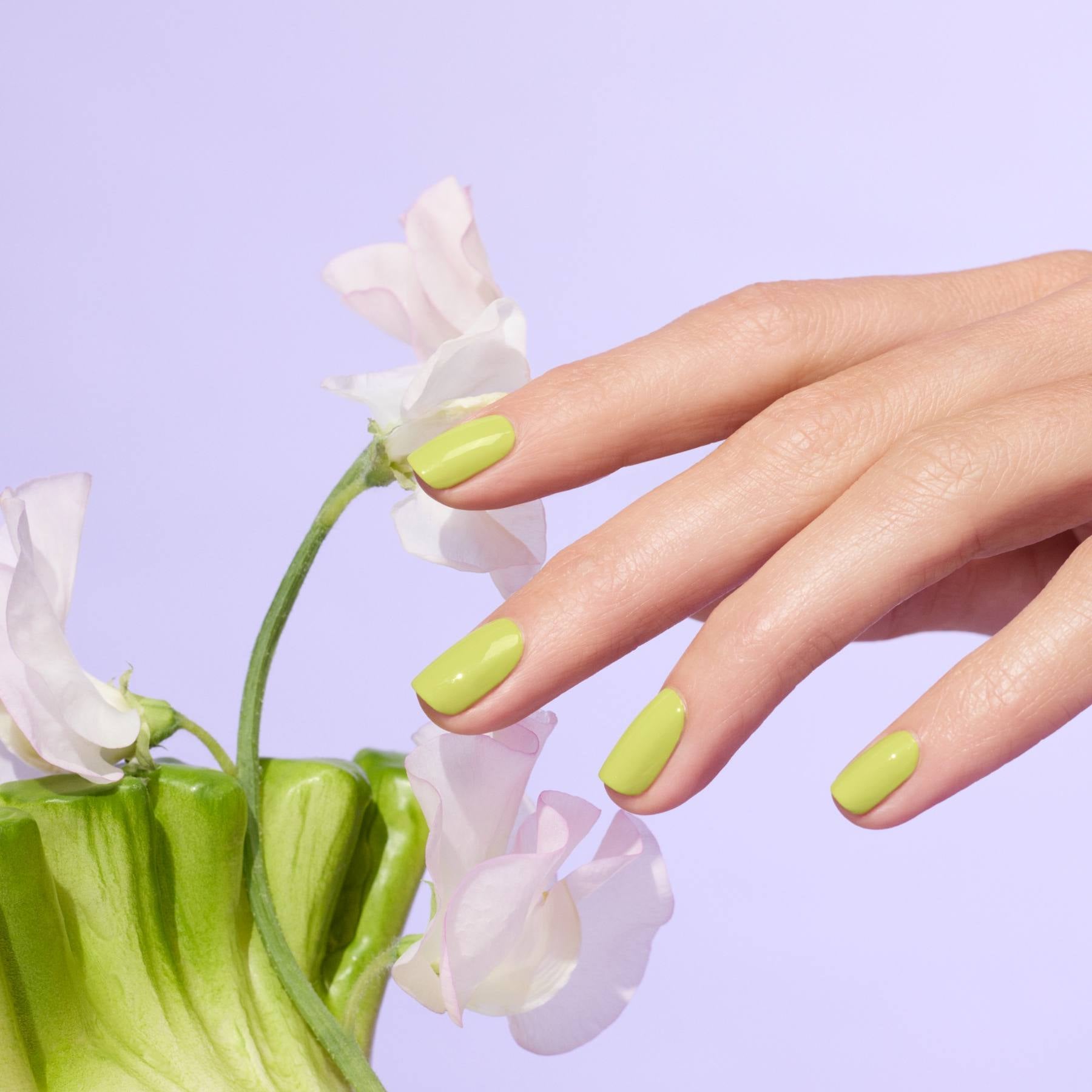 A hand with Manucurist Lime gel polish manicure touches delicate pale pink flowers against a pastel purple background.