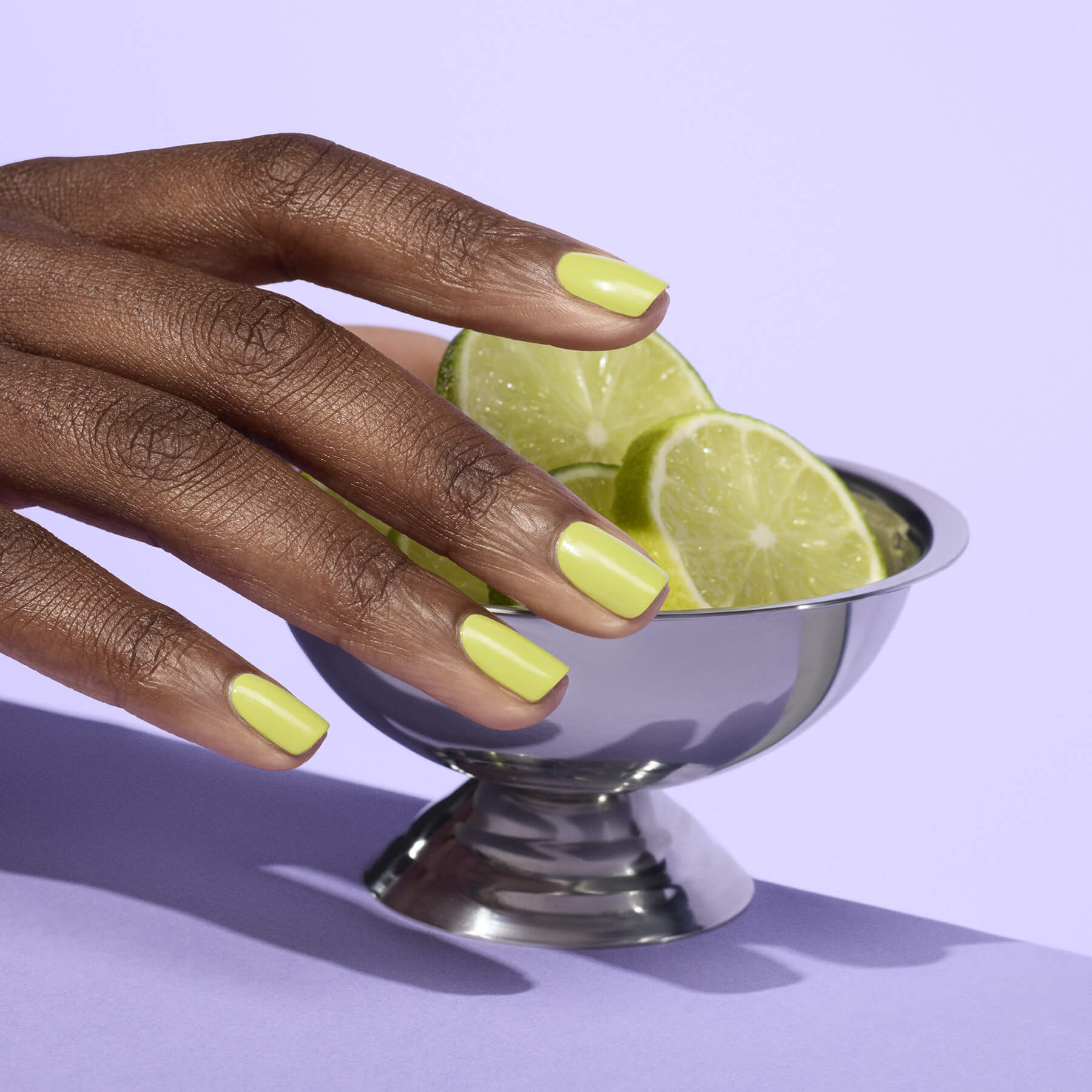 A hand with yellow-green nail polish rests on the edge of a silver bowl filled with lime slices, set against a light purple background.