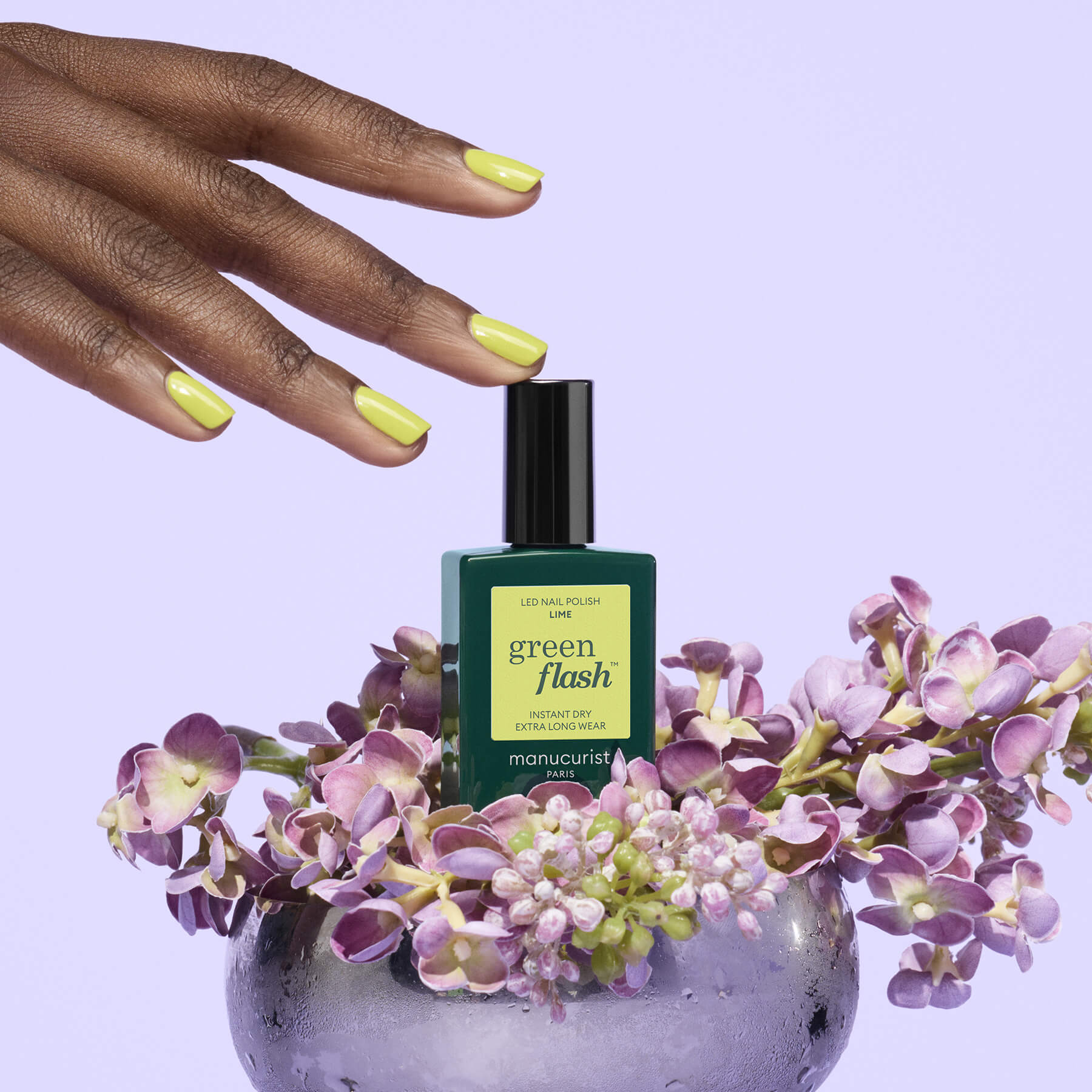 A hand with yellow-green painted nails hovers above a chartreuse Manucurist Lime gel polish bottle, surrounded by purple flowers in a clear vase, set against a pale lavender background.