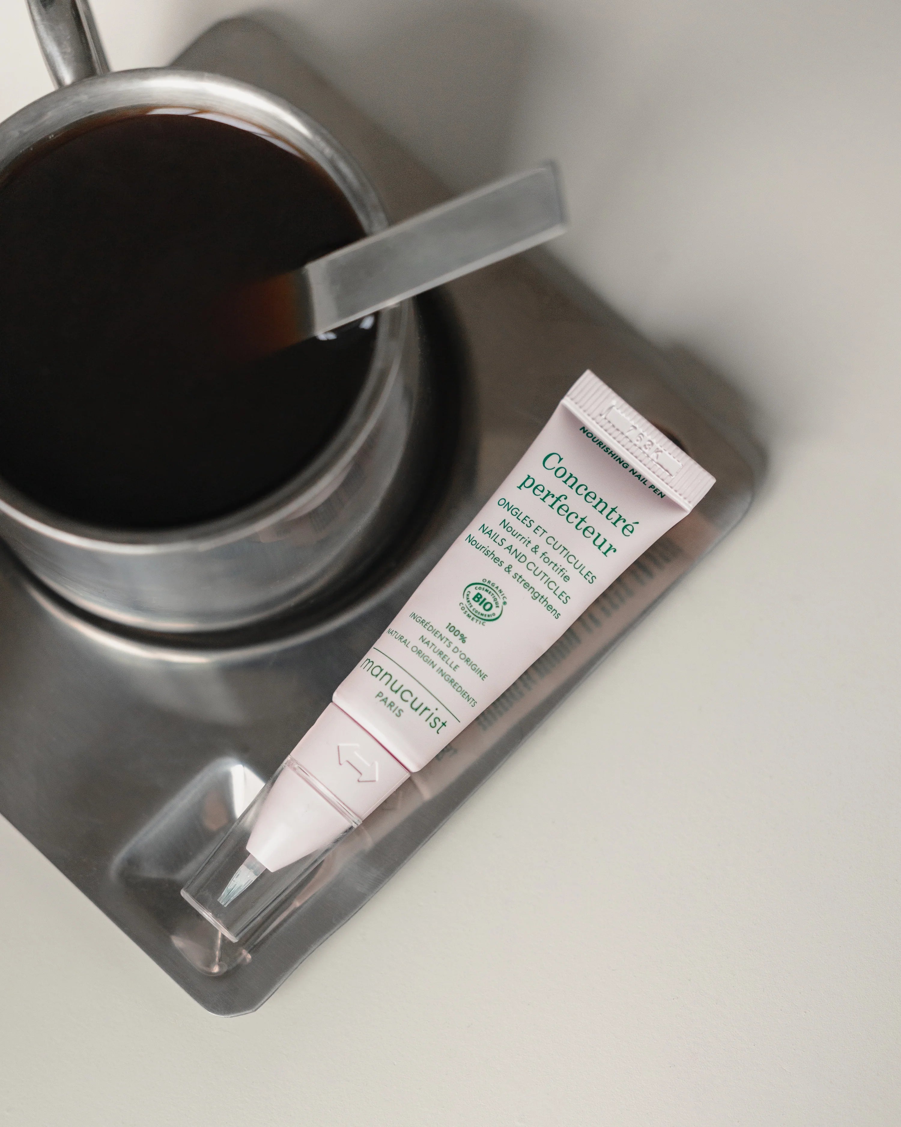 A tube of Concentré perfecteur face care lies on a metal tray next to a cup of black coffee with a metal handle, set on a light-colored surface.