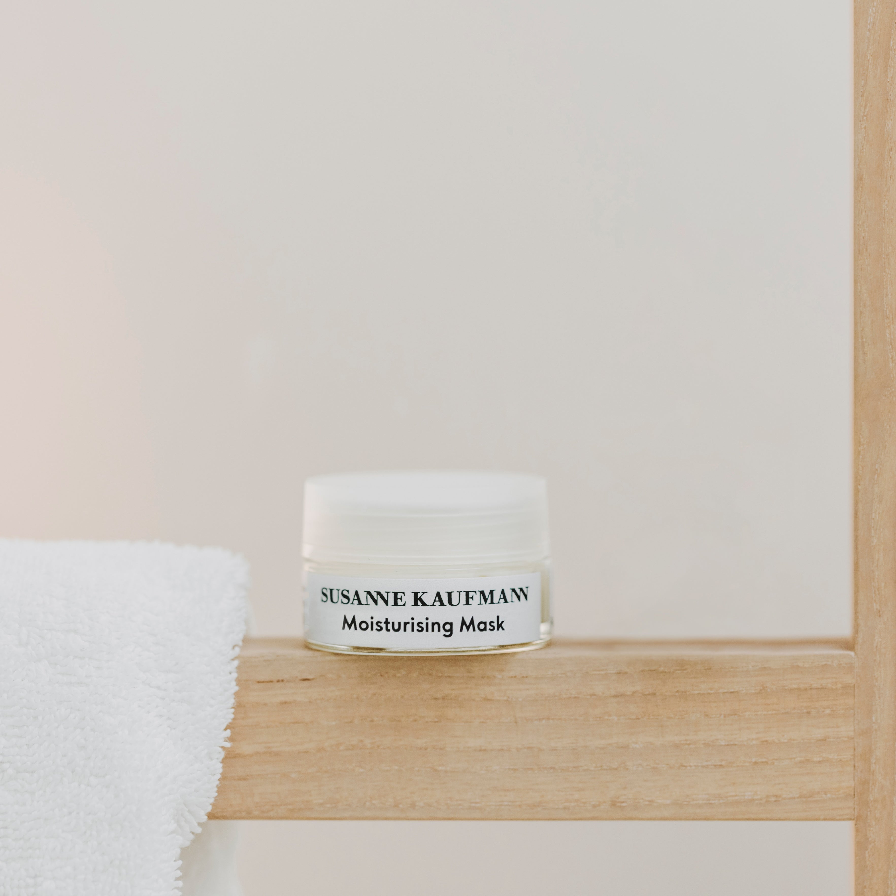 A small white jar labeled SUSANNE KAUFMANN Moisturising Mask sits on a light wooden shelf next to a neatly folded white towel against a plain background.