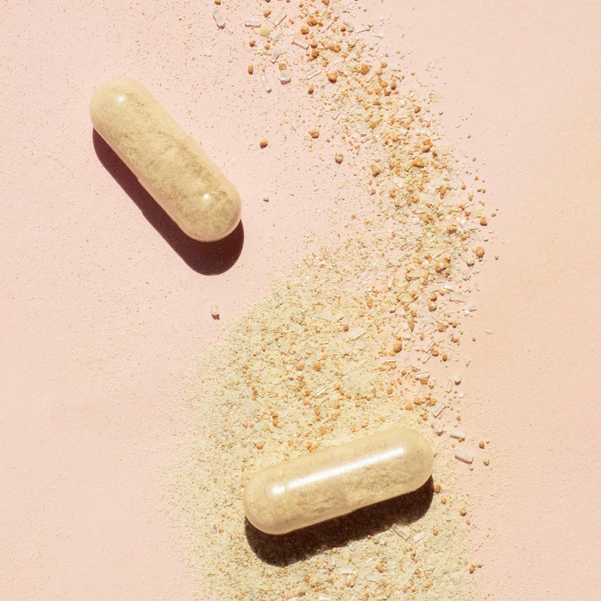 Deux capsules Nail Boost beiges de Manucurist reposent sur une surface rose clair, recouvrant en partie de la poudre beige éparpillée — idéales pour celles et ceux qui recherchent des compléments pour renforcer les ongles et favoriser une croissance saine des ongles.