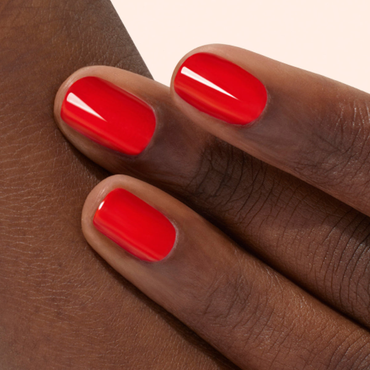 Gros plan sur trois doigts à la peau foncée, avec des ongles soigneusement manucurés et vernis d’un rouge vif brillant. Les doigts reposent délicatement sur le dos d’une main.