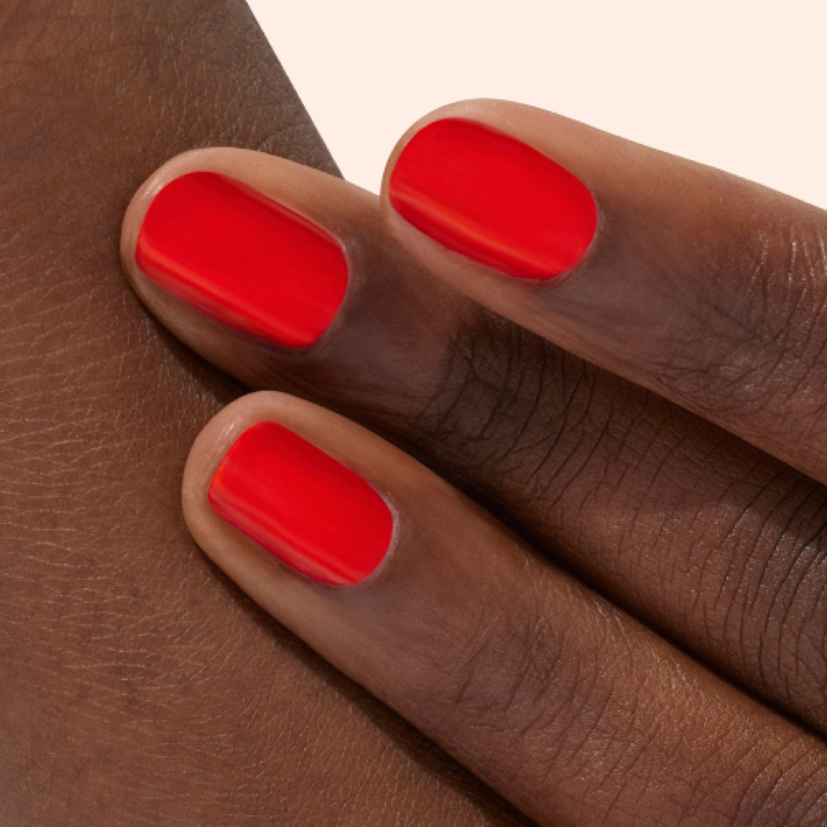 Gros plan sur trois doigts à la peau foncée et lisse, arborant des ongles impeccablement manucurés et vernis d'un rouge vif et brillant. L'arrière-plan est d'une couleur douce et pâle.