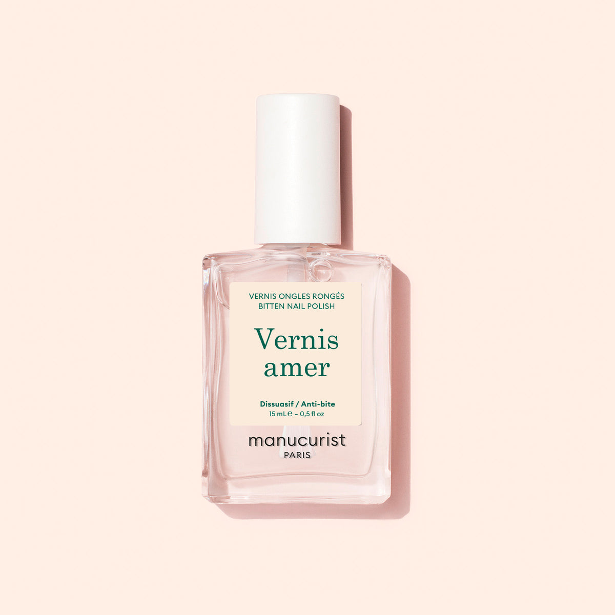 Un flacon rectangulaire transparent de Vernis Amer Manucurist au bouchon blanc repose sur un fond rose pâle. Son étiquette crème au texte vert et beige met en avant des bienfaits de soin des ongles, aidant à lutter contre le rongement des ongles.