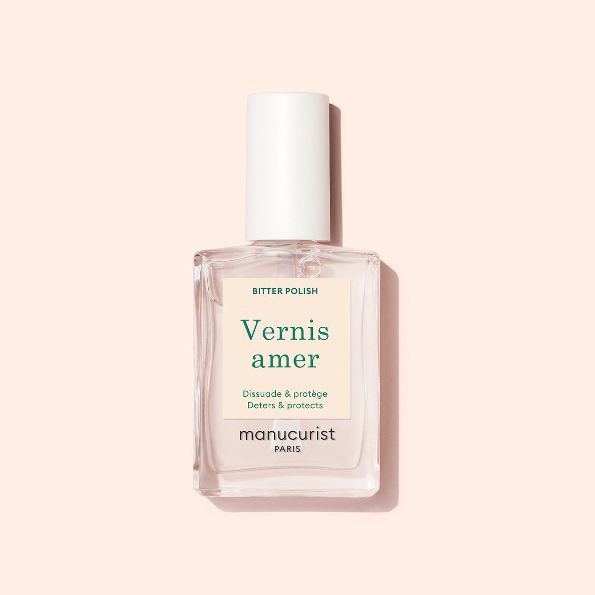 Un flacon rectangulaire transparent de Vernis Amer Manucurist au bouchon blanc repose sur un fond rose pâle. Son étiquette crème au texte vert et beige met en avant des bienfaits de soin des ongles, aidant à lutter contre le rongement des ongles.