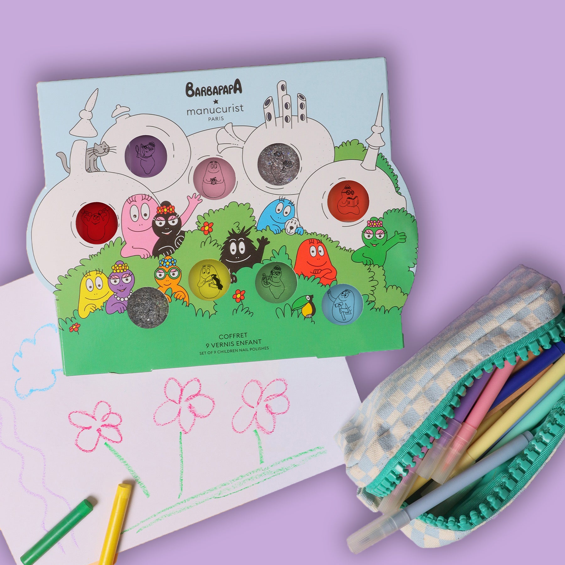 A colorful Barbapapa-themed nail polish set is displayed on a light purple surface next to a pencil case and a drawing of flowers and clouds made with colored pencils.