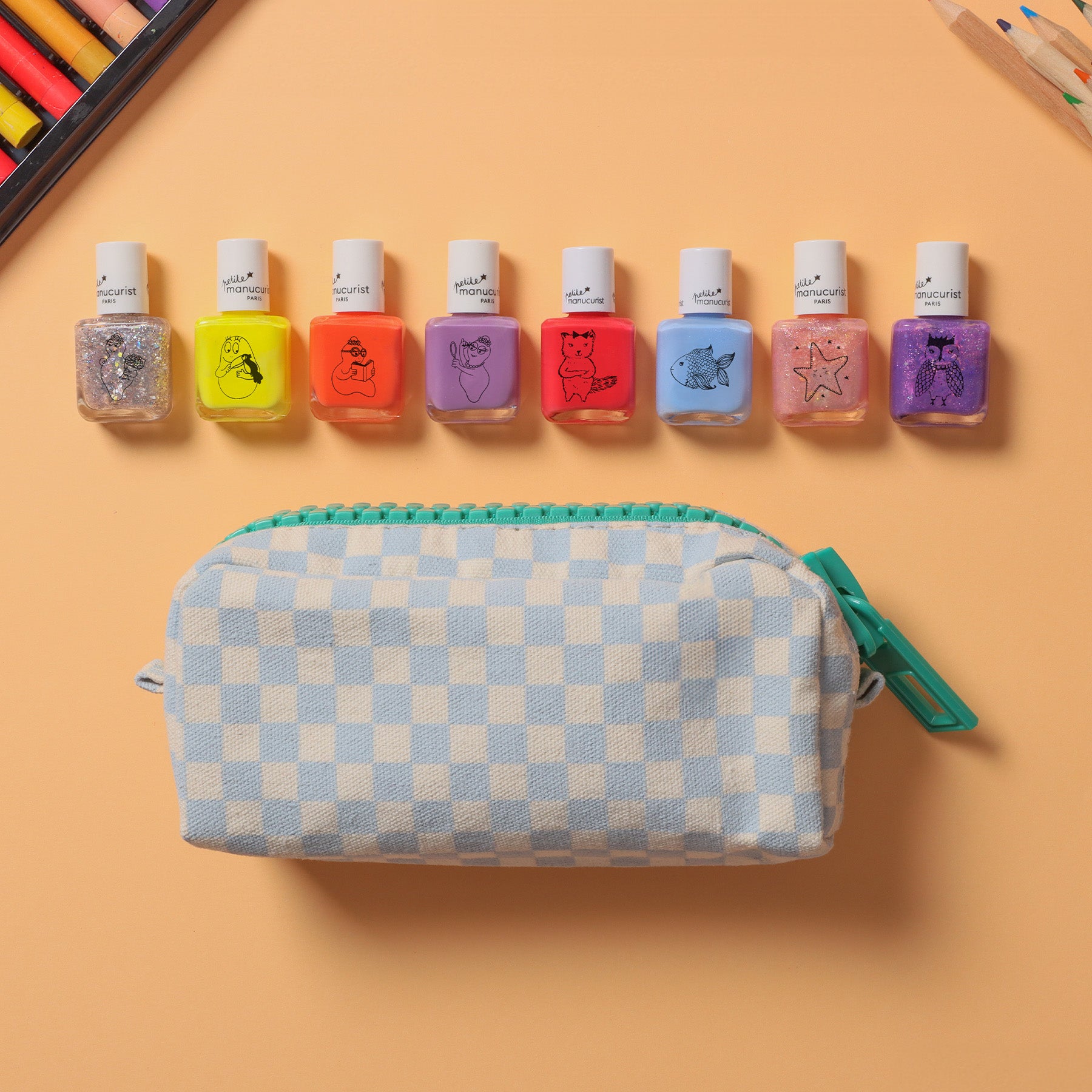 A checkered white and light blue zippered pouch sits below seven bottles of colorful nail polish, each with a different cartoon design, arranged in a row on a peach background. Colored pencils are partially visible in the corner.