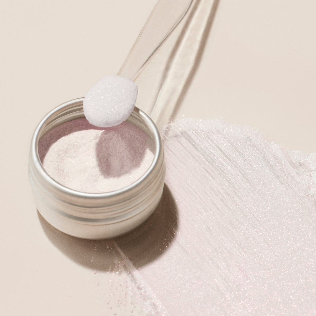 Un petit pot en métal de poudre effet glacé rose Manucurist, rempli d’une fine poudre blanche au subtil éclat nacré, est posé sur une surface claire. Une cuillère transparente en plastique contient un peu de poudre scintillante à côté du pot.