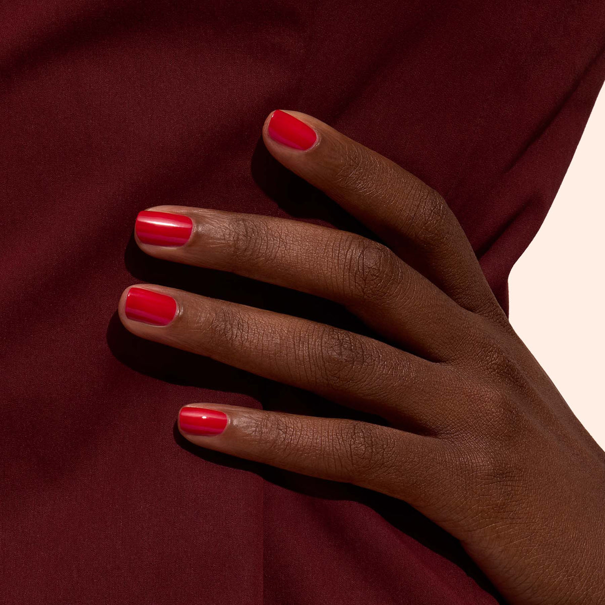Une main à la peau foncée aux ongles soigneusement manucurés, vernis en Poppy Red de Manucurist, repose sur un fond en tissu bordeaux.