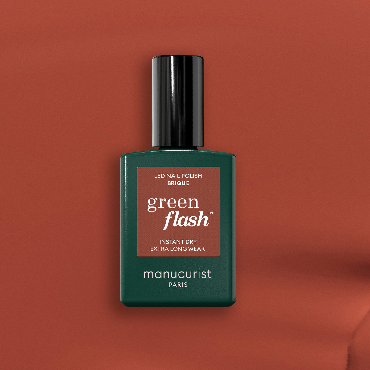 Un flacon de vernis à ongles LED Green Flash Manucurist Brique sur un fond marron chaud. Le flacon vert foncé au bouchon noir et à l&