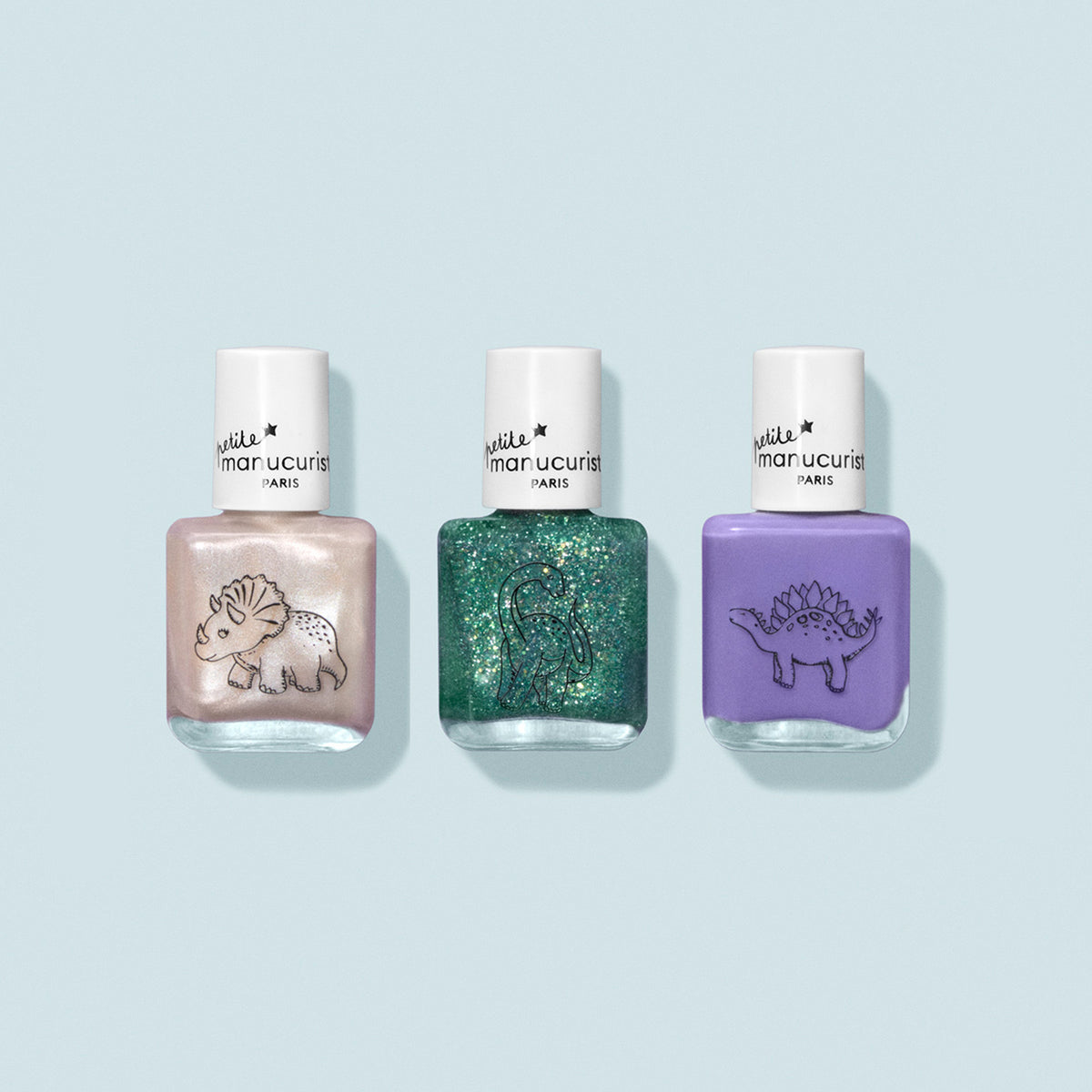 The Manucurist Wild Life Collection kids nail polish set includes three gentle-formula bottles: pearly white with a triceratops, glittery green with a brontosaurus, and pastel purple with a stegosaurus, all on a pale blue background.