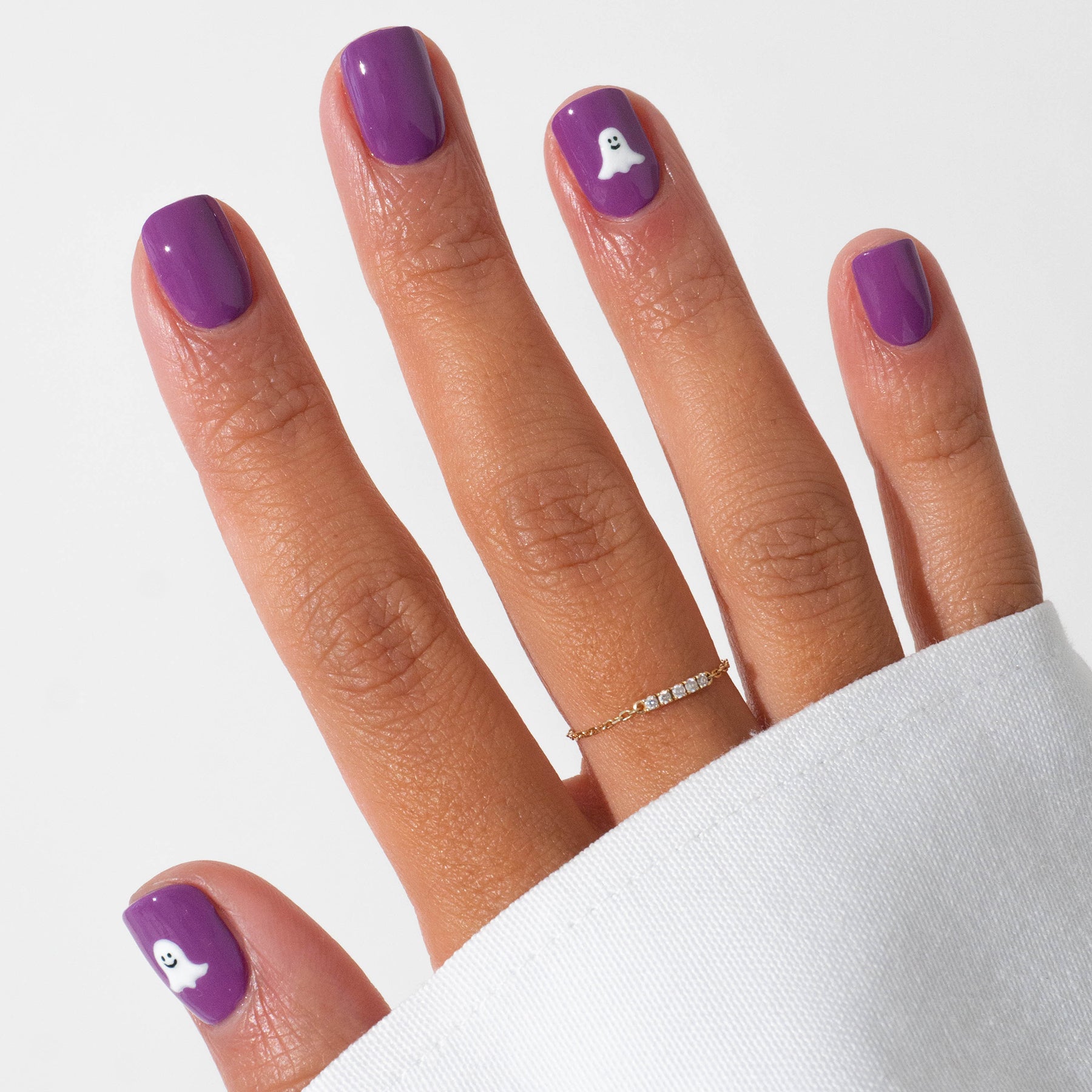 A hand sports Manucurist’s Witchy Wicked purple gel polish, with two nails adorned with white ghost Halloween art. A thin gold ring is worn, and a white sleeve partially covers the hand.