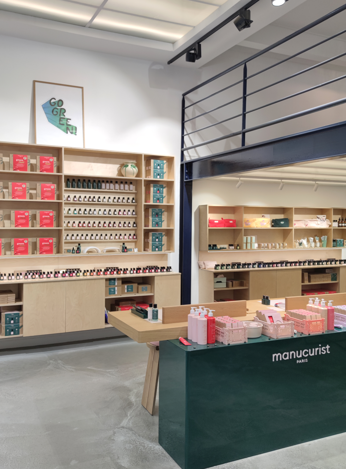 A modern nail salon with shelves of nail polish, skincare products, and a green counter labeled manucurist paris. A Go Green poster hangs on the wall above the shelves.