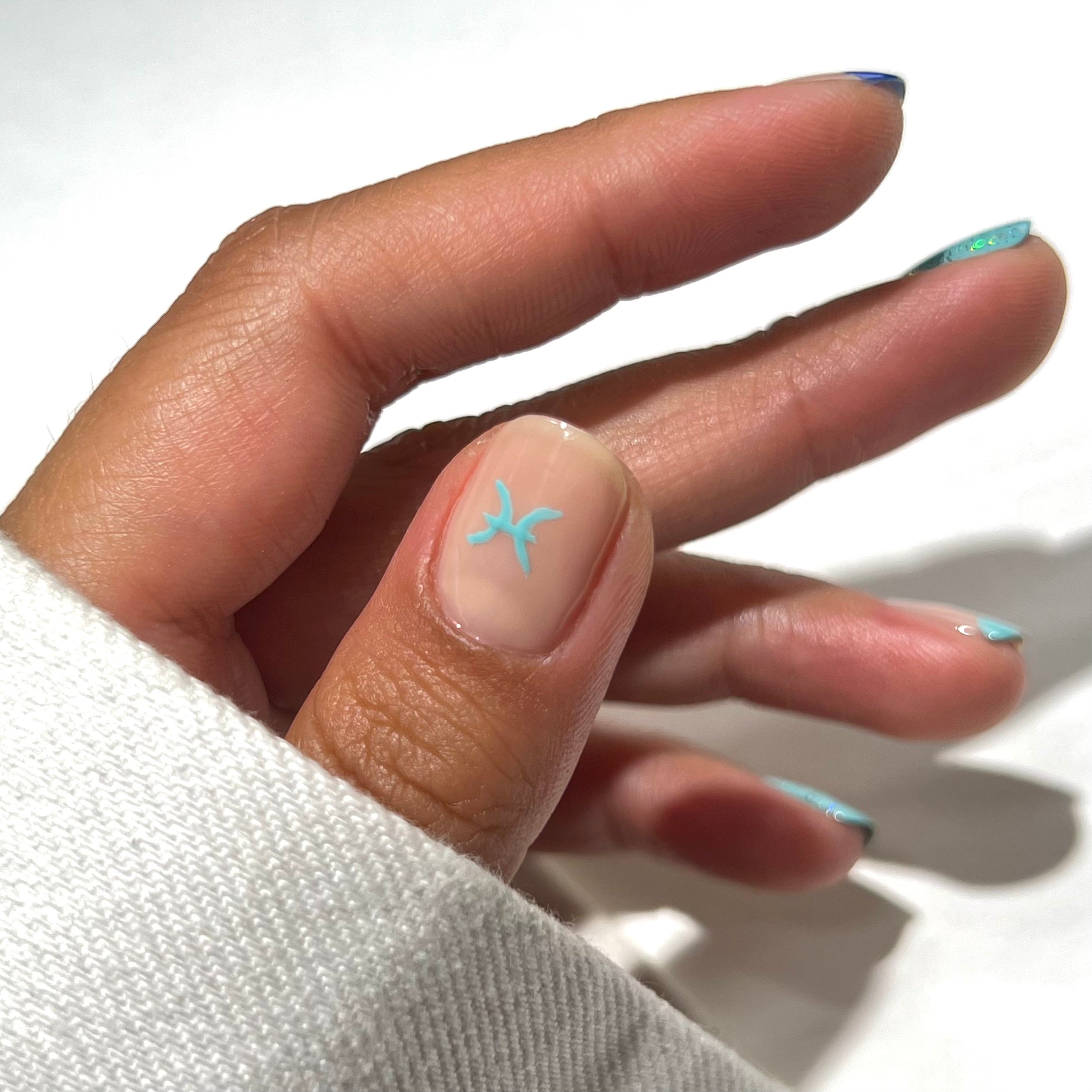 A hand with a light brown skin tone, wearing a white sleeve. The thumbnail has a small turquoise Pisces zodiac symbol, and the other nails have turquoise tips with minimal designs. The background is white.