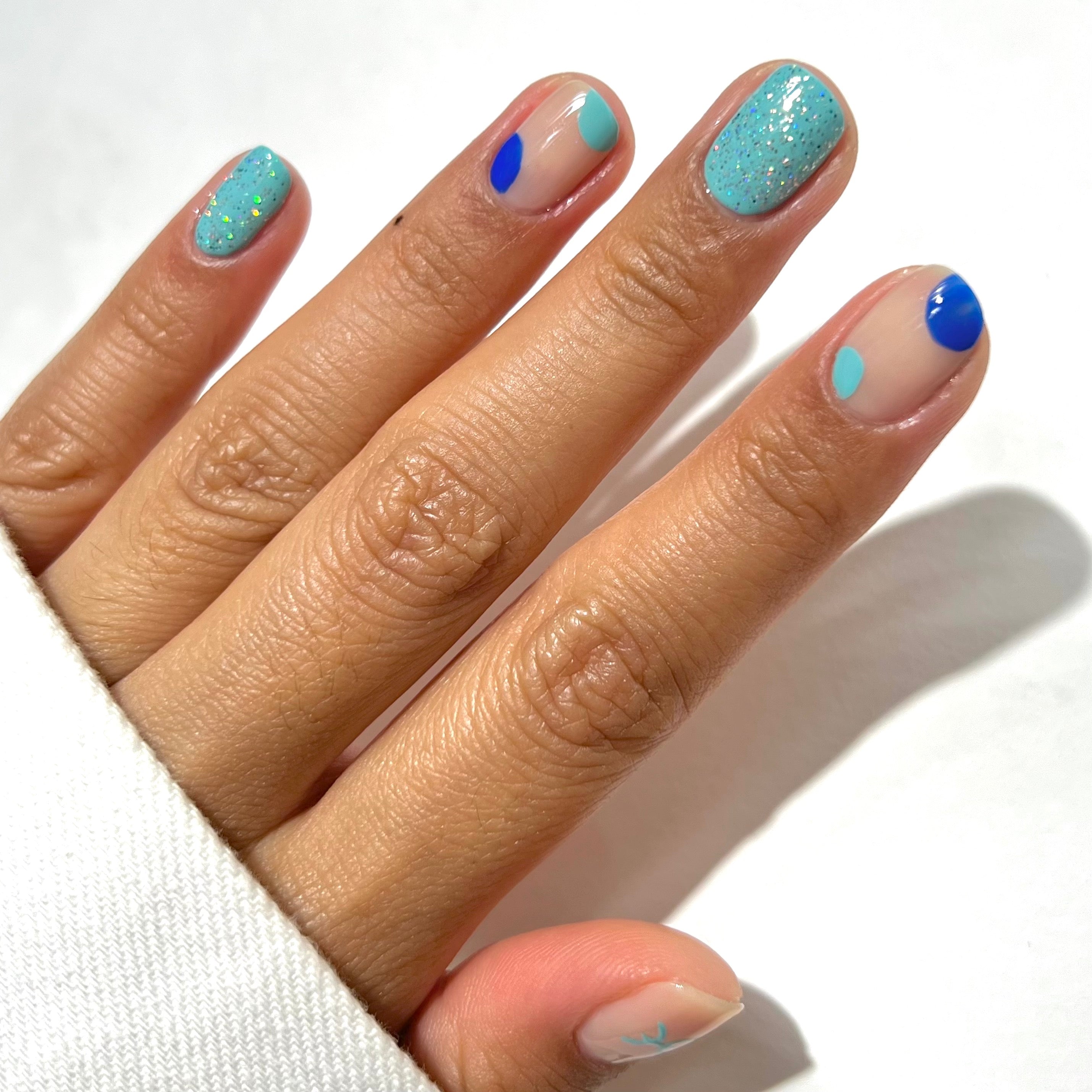 A hand with a white sleeve showing nails painted in blue and teal designs, including solid, glitter, and abstract dots on a light background.