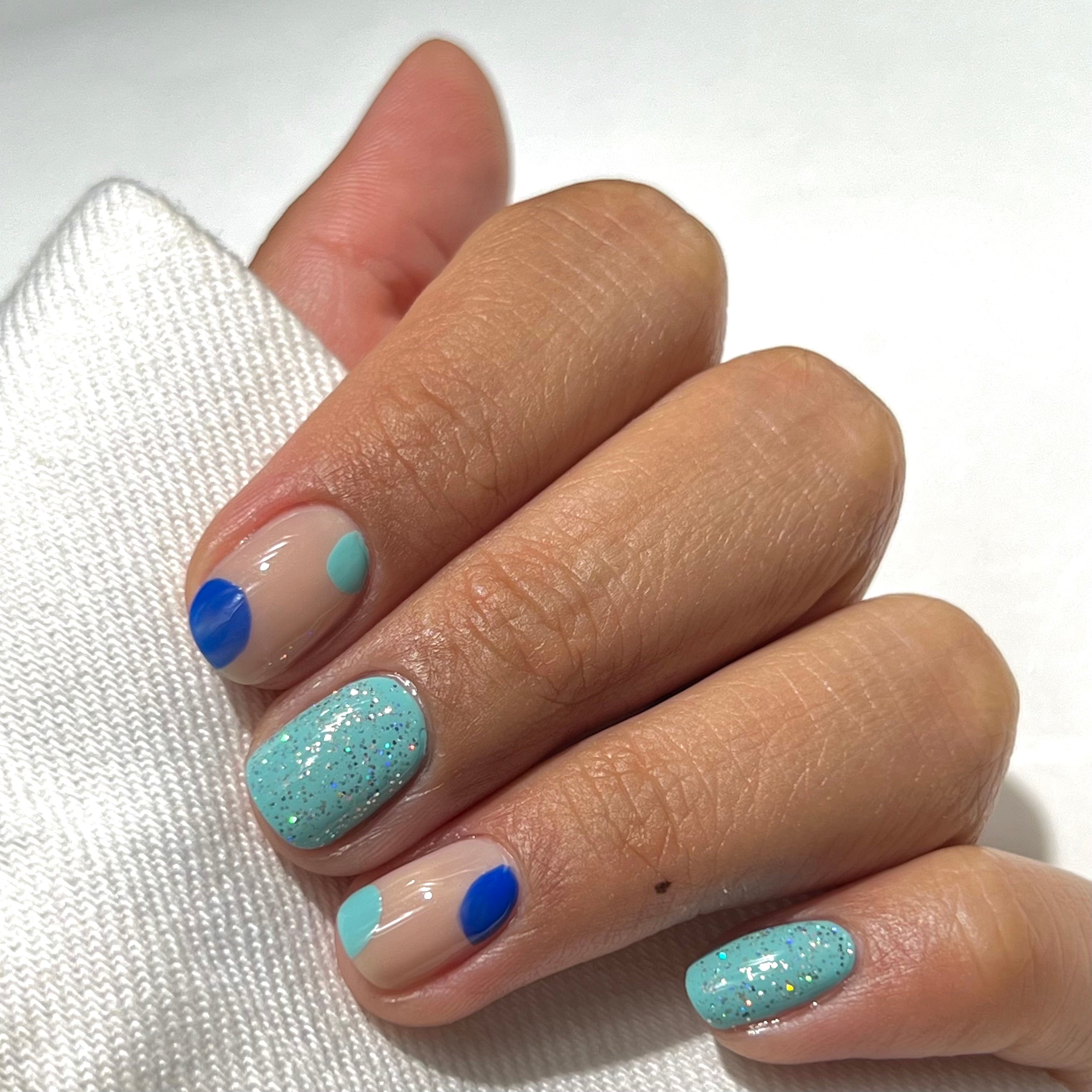 A hand with short nails features blue and teal dots on a nude base, with two nails painted light teal and accented with glitter. The hand holds a white fabric.