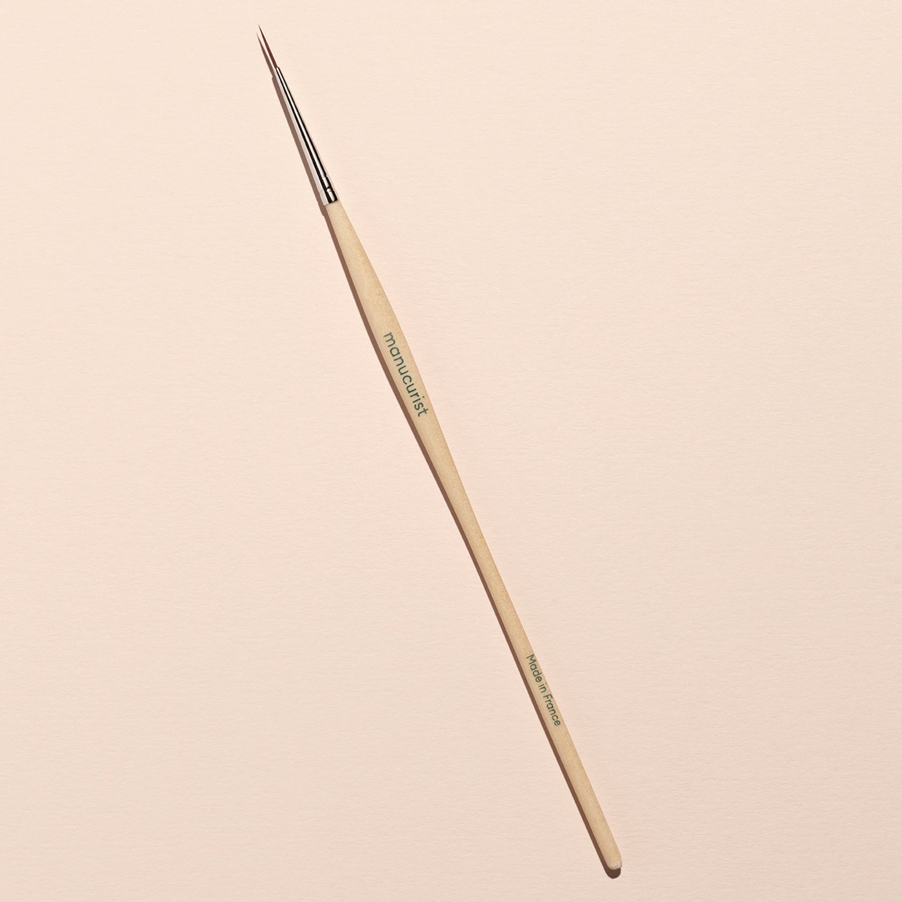 The Witchy Wicked brush by Manucurist, featuring a wooden handle and metal ferrule, lies diagonally on a light beige surface—ideal for detailed Halloween nail art with gel polishes or adding biodegradable glitter accents.