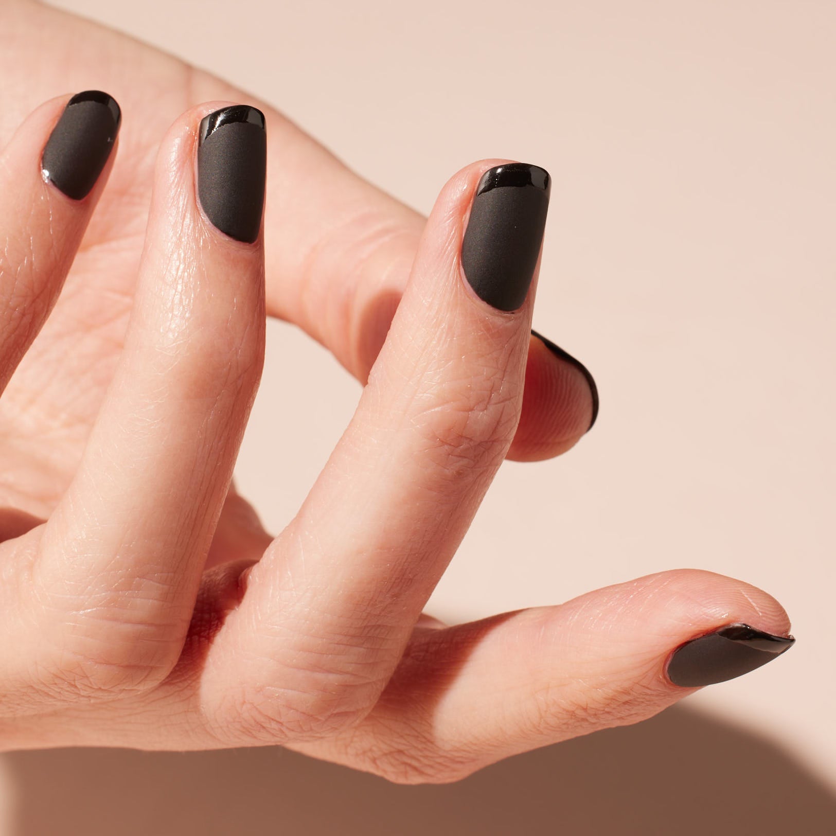 A close-up of a hand with fingers slightly curled, showing nails painted matte black with glossy black tips, against a soft beige background.