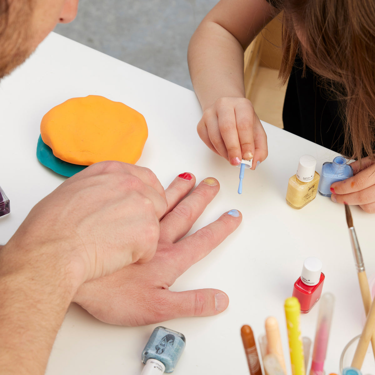 A child paints an adult’s nails with Manucurist’s You Me Under the Sea Green Flash™ Set at a white table, surrounded by bottles of polish, art supplies, and modeling clay.