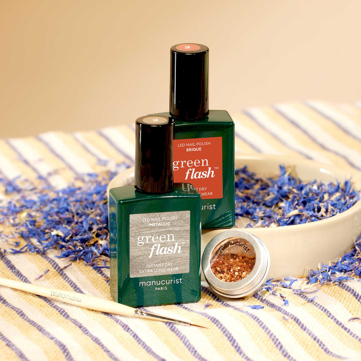 Deux flacons de vernis à ongles Manucurist Oriental Vibe et un pot de paillettes biodégradables dorées de la collection Automne 2023 sont disposés dans un bol beige sur un tissu rayé, entourés de pétales de fleurs séchées bleues et violettes. Un pinceau de manucure est posé à proximité.