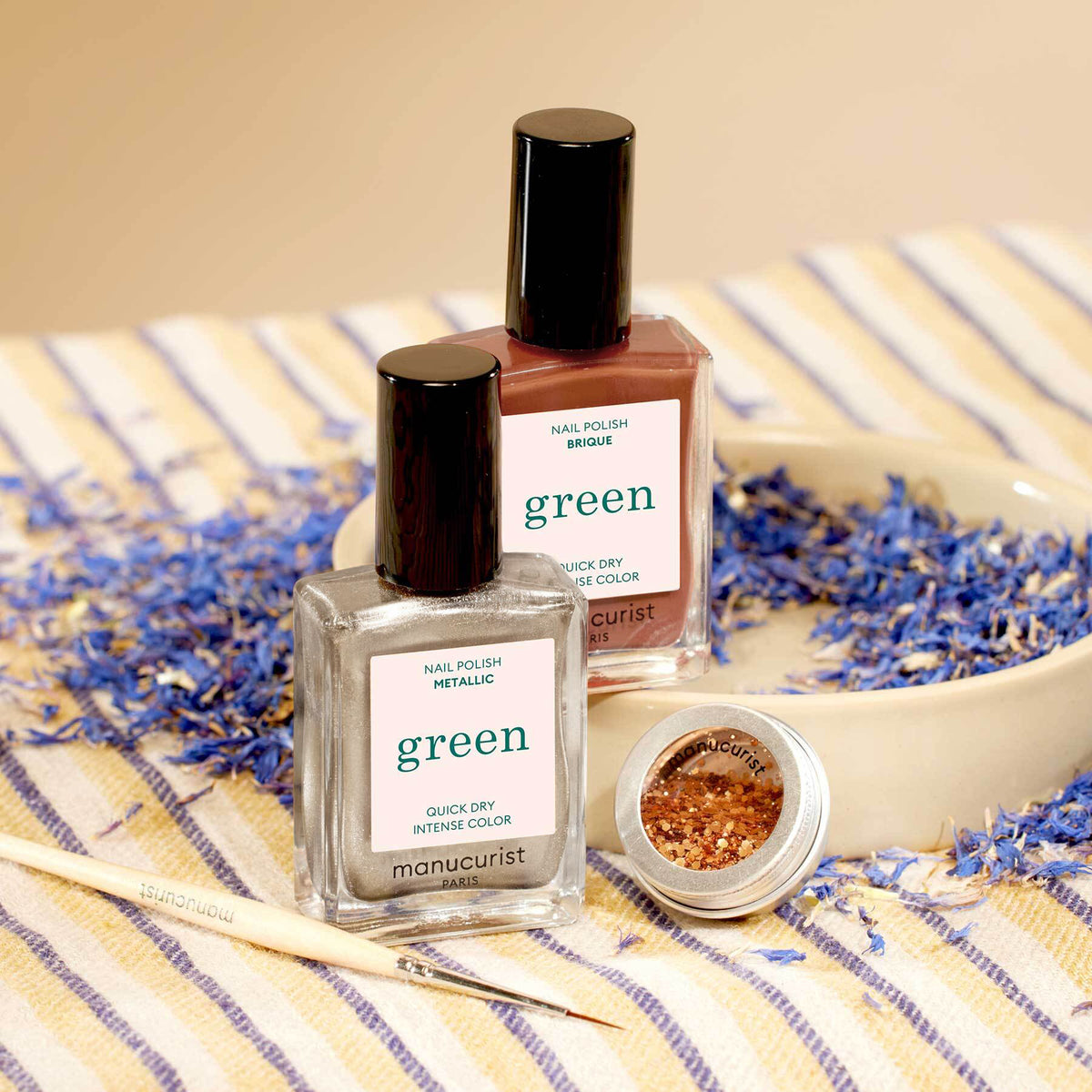 Deux flacons de vernis à ongles Manucurist Oriental Vibe (argent métallisé et rouge brique) de la collection Automne 2023 et un petit pot ouvert de paillettes biodégradables reposent sur une assiette avec des pétales bleus séchés, sur un tissu rayé. Un pinceau est posé à côté.