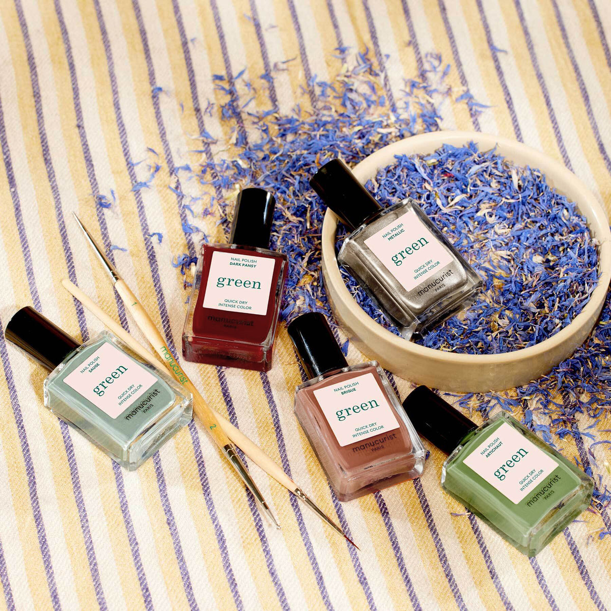 Cinq flacons de vernis à ongles Green Flash Manucurist de la collection Automne 2023 sont présentés sur un tissu rayé, avec des fleurs séchées bleues, un bol en bois et deux outils de Nail Art Manucurist Spice Lover à proximité.