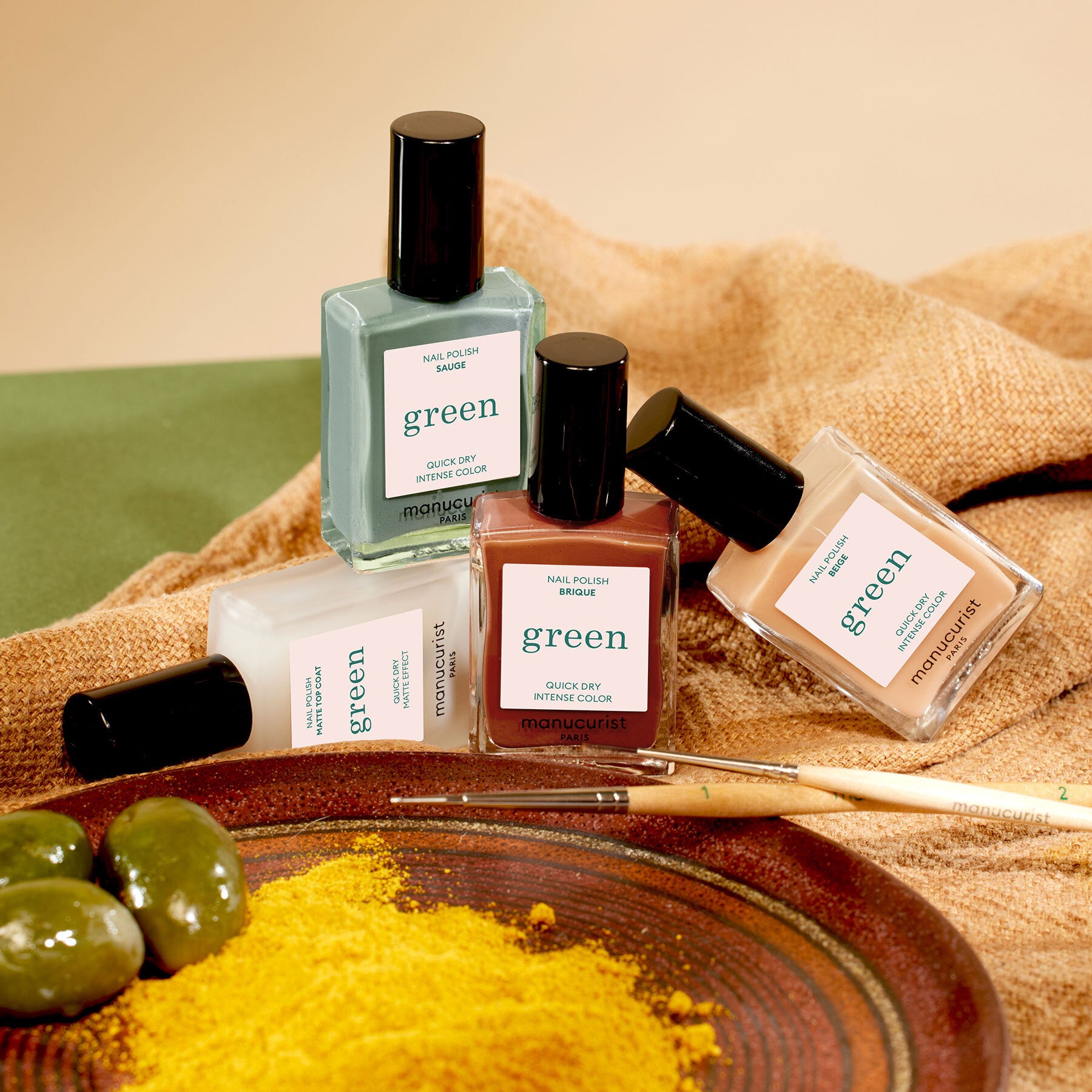 Four bottles of Manucurist’s Geometric Obsession nail polish are arranged on a brown cloth with paintbrushes, green olives, and yellow powder, creating an inspiring setup for modern nail art.