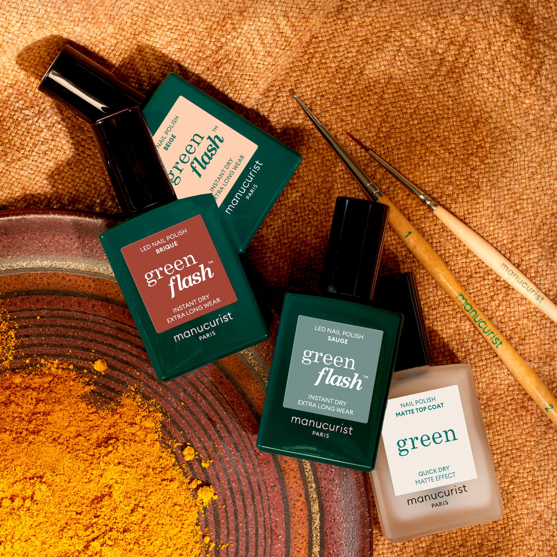 Four bottles of Manucurist Geometric Obsession nail polish in green and cream shades from the Autumn 2023 collection are displayed on a textured brown surface with yellow powder, two nail art brushes, and geometric nail art inspiration.