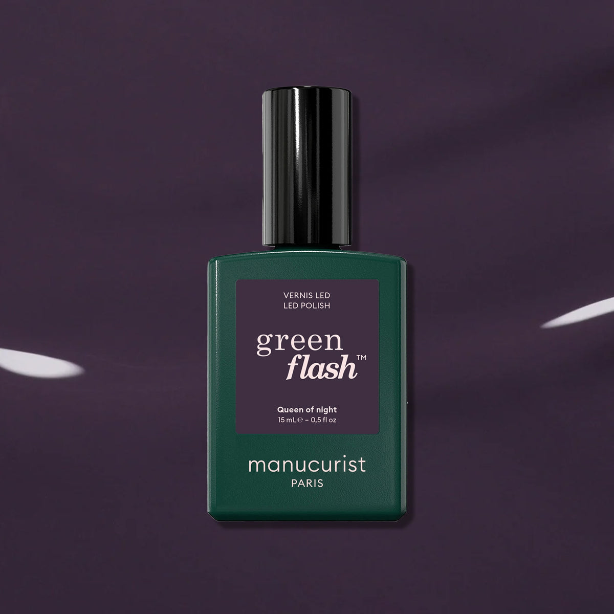 A dark green Manucurist Queen of Night nail polish bottle rests against a glossy purple backdrop, highlighting its gray undertones and unique style.