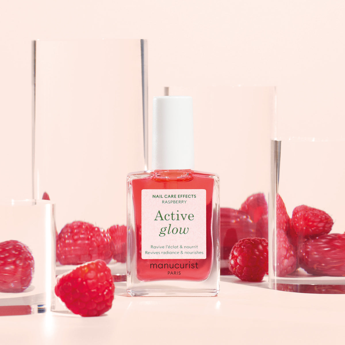 Un flacon de soin pour les ongles Manucurist Active™ Glow Raspberry repose parmi des blocs d’acrylique transparent et des framboises fraîches sur un fond rose. L’étiquette met en avant sa formule d’origine végétale, pour des ongles éclatants et un soin nourrissant infusé des bienfaits fruités de la framboise.