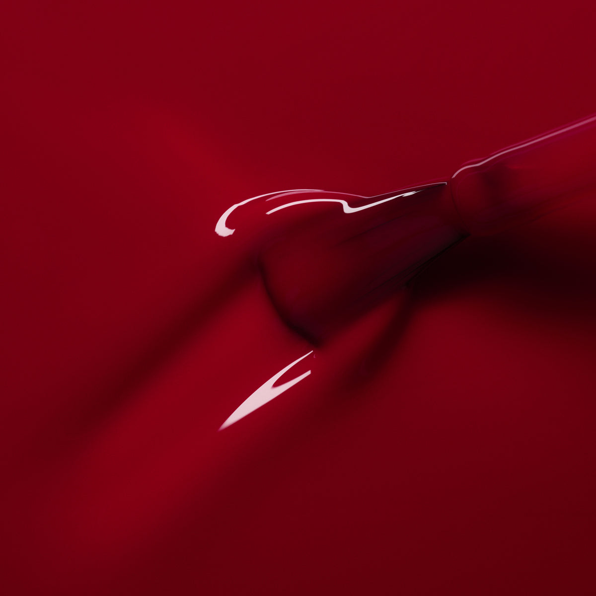 A close-up of a dropper releasing Manucurist’s Pomegranate nail polish onto a glossy red surface, forming a smooth, reflective texture.
