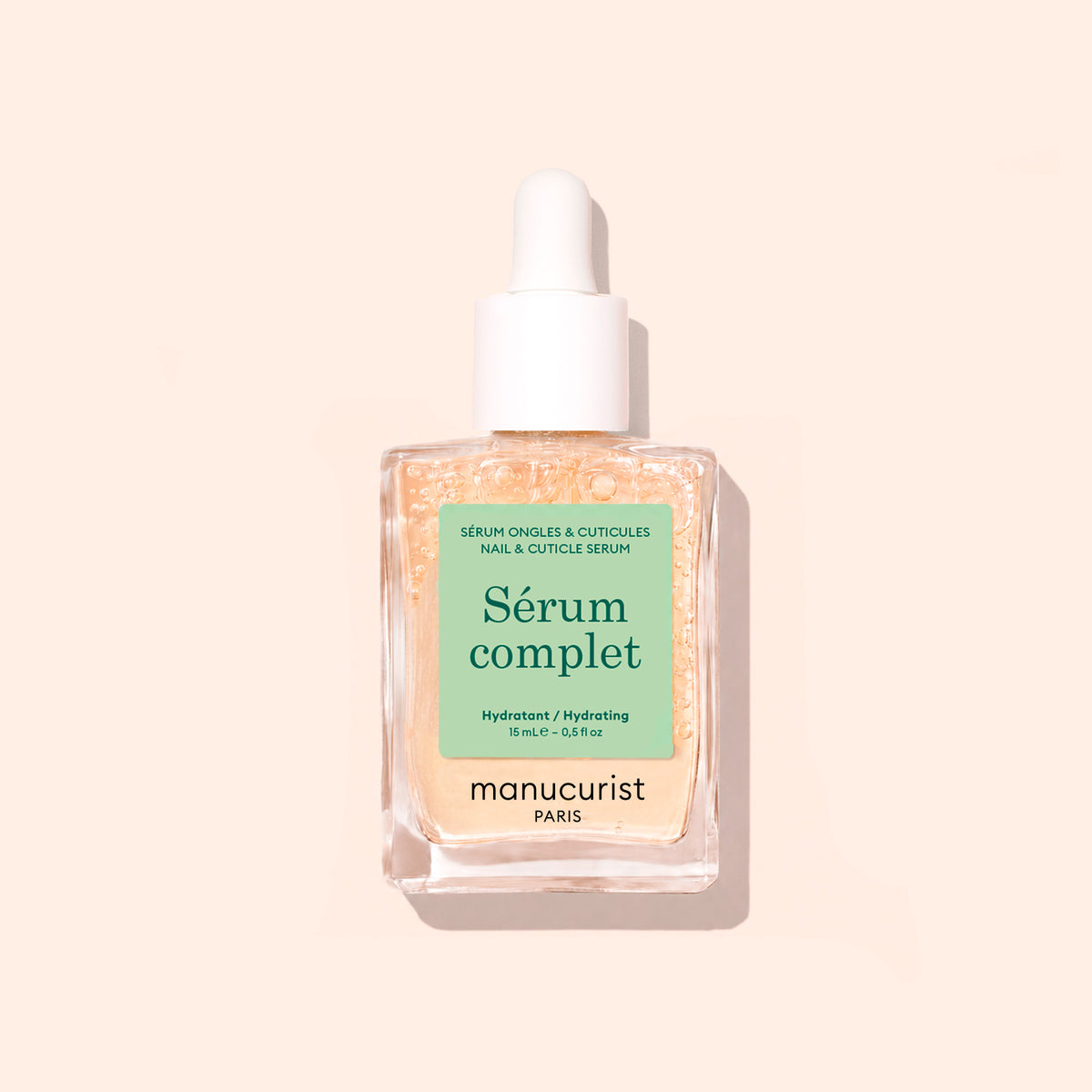 Un flacon compte-gouttes en verre transparent étiqueté Nail Hydrating Serum par Manucurist, contenant un sérum nourrissant pour les ongles au liquide beige clair, présenté sur un fond beige clair.