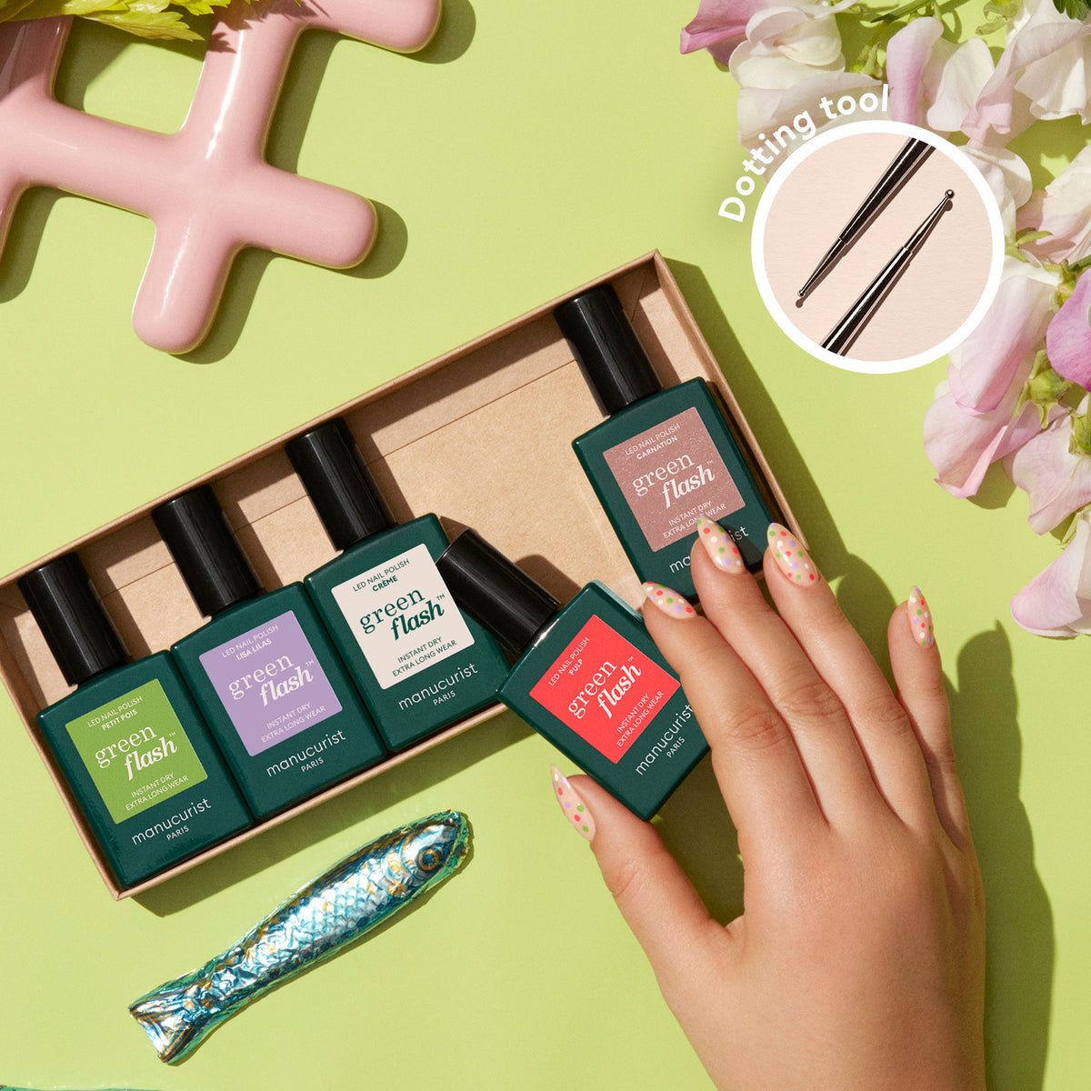 Une main aux ongles décorés tient un Manucurist Dot it better! près d’une boîte de cinq flacons, entourée de fleurs, d’une figurine de poisson et d’un objet rose. Un encart met en avant l’outil Dot it better! à double embout pour un Nail Art créatif.