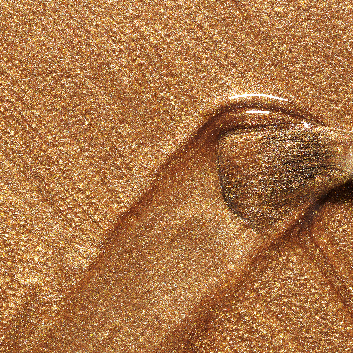 Un gros plan d’un pinceau appliquant Shimmer de Manucurist, étalant un vernis à ongles bronze au fini glacé au miel et formant une traînée brillante sur une surface dorée et scintillante.